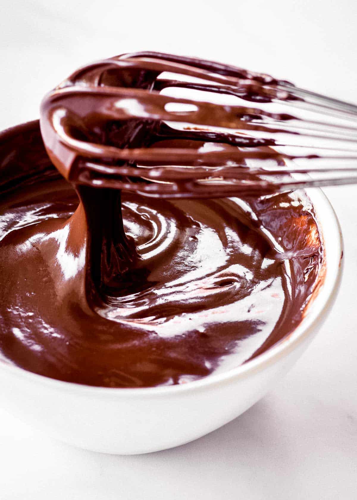 Vegan chocolate ganache drips off a whisk and into a white bowl.
