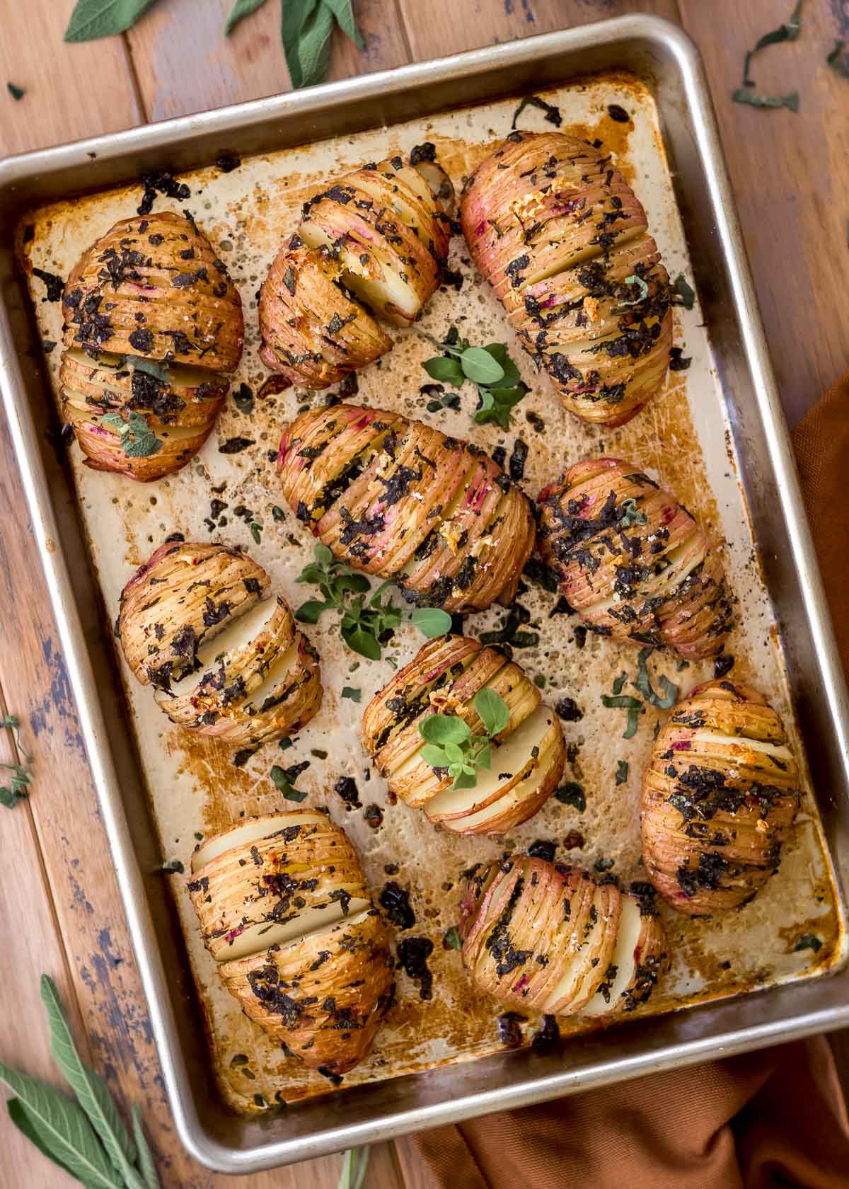 Garlic hasselback potatoes on a silver baking sheet, decorated with fresh herbs.