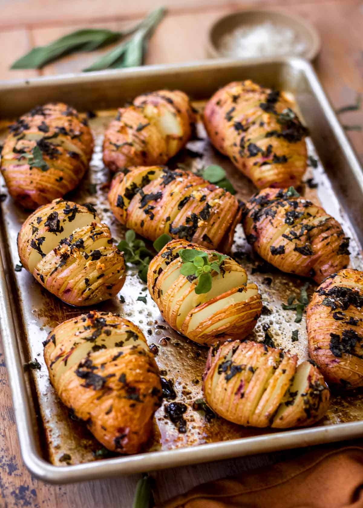 Garlic hasselback potatoes on a silver baking sheet, decorated with fresh herbs and herbs in the background.