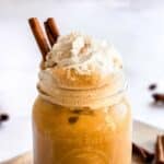 Iced pumpkin spice latte in a glass, topped with coconut whipped cream, cinnamon sticks and pumpkin spice. Extra cinnamon sticks and star anise are in background.