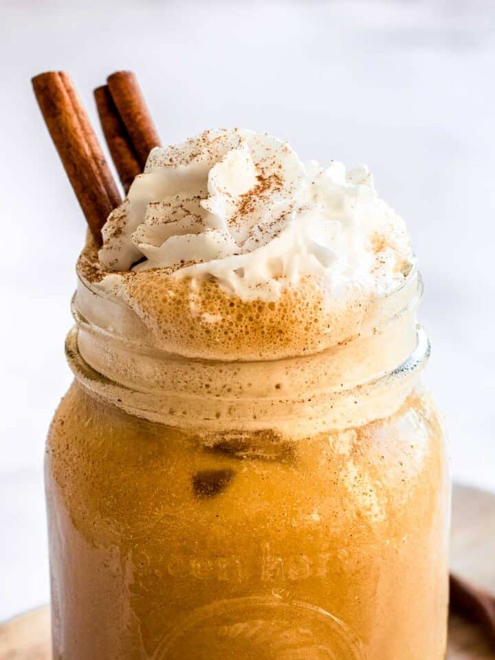 Iced pumpkin spice latte in a glass, topped with coconut whipped cream, cinnamon sticks and pumpkin spice. Extra cinnamon sticks and star anise are in background.