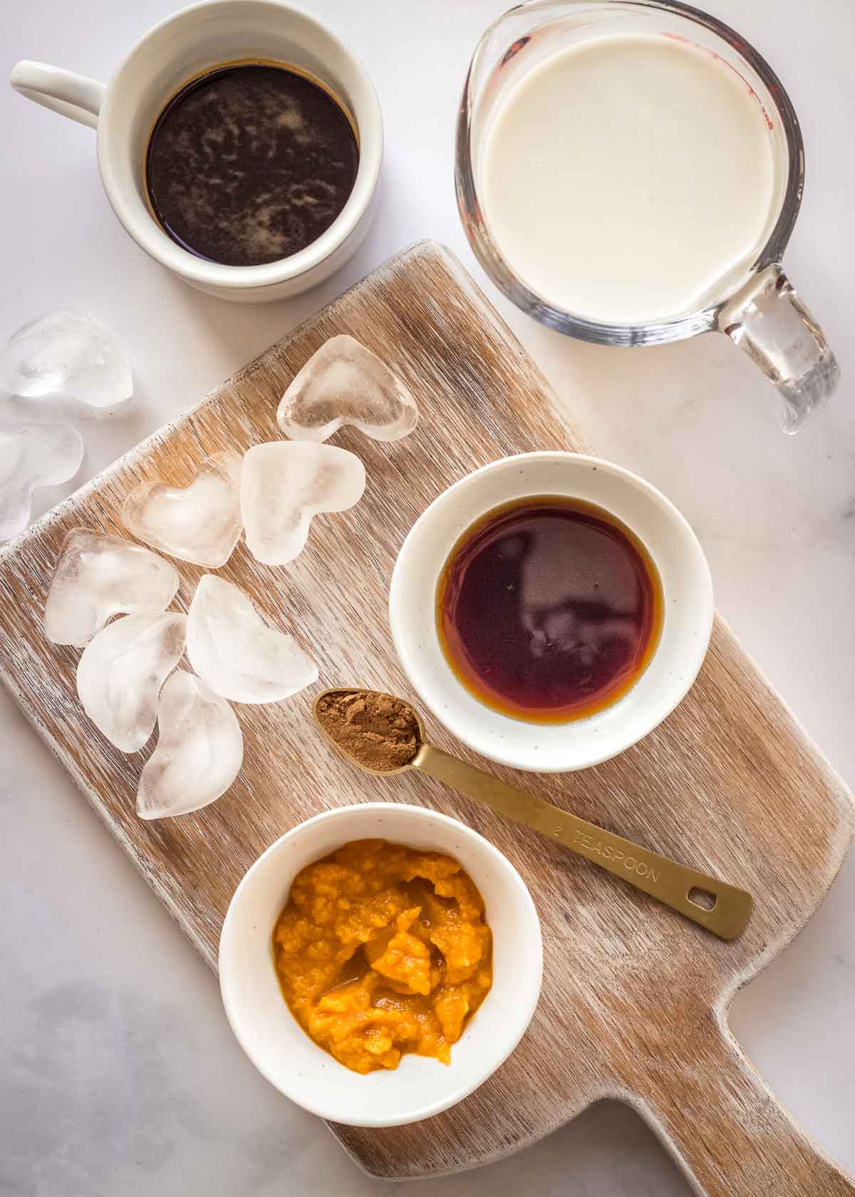 Ingredients for pumpkin spice latte in bowls on a chopping board, including ice, pumpkin puree and milk.