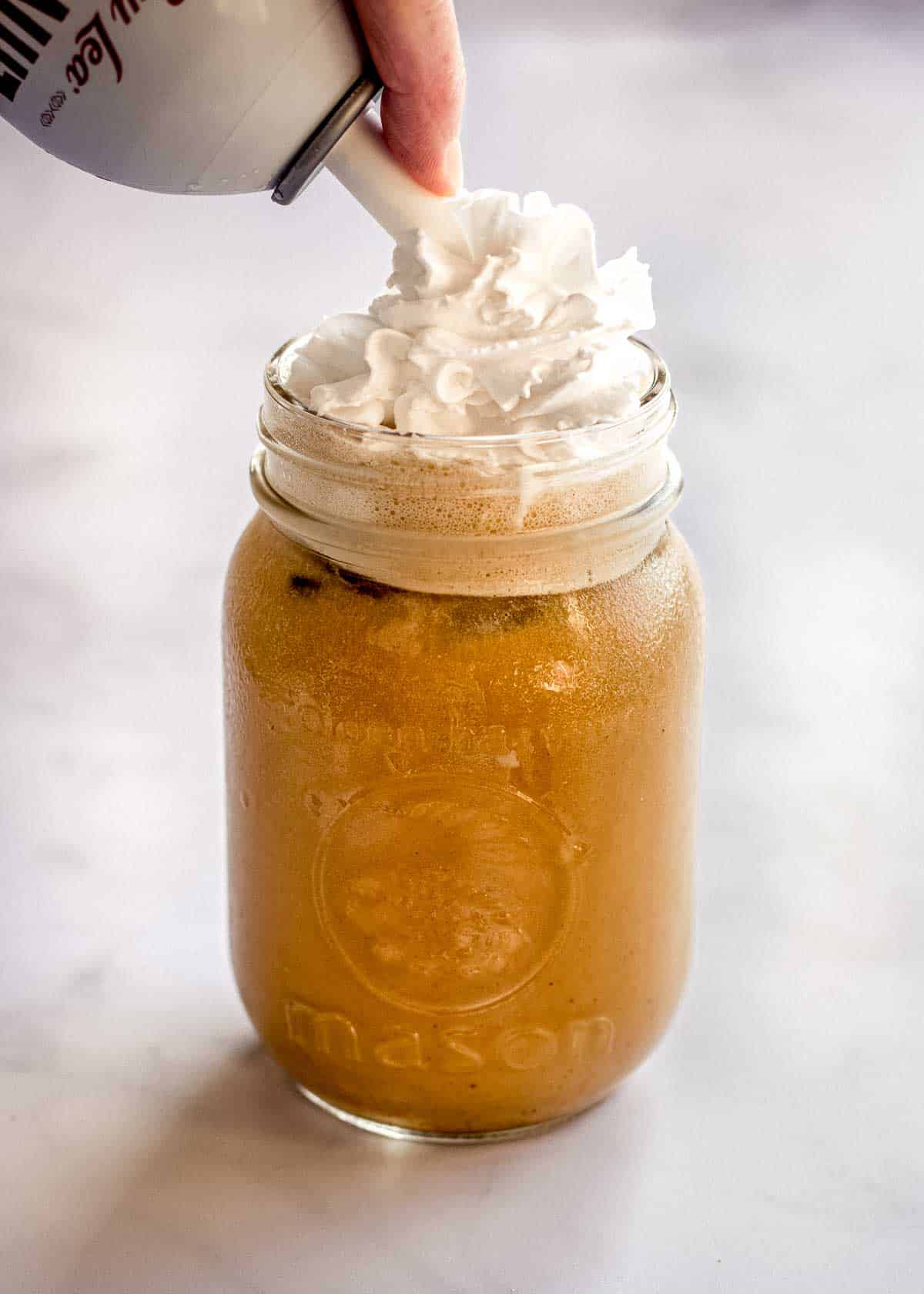 A woman's hand decorates iced pumpkin spiced latte in a glass with whipped cream from a can.
