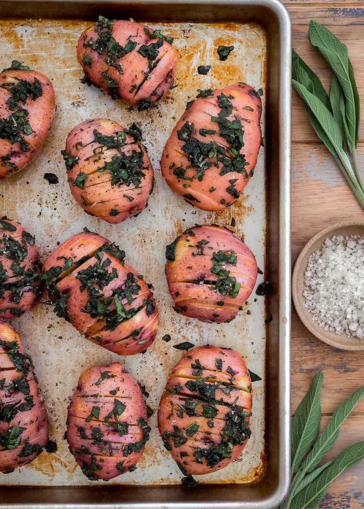 Sage-filled hasselback red potatoes on a silver baking sheet with sage leaves nearby.