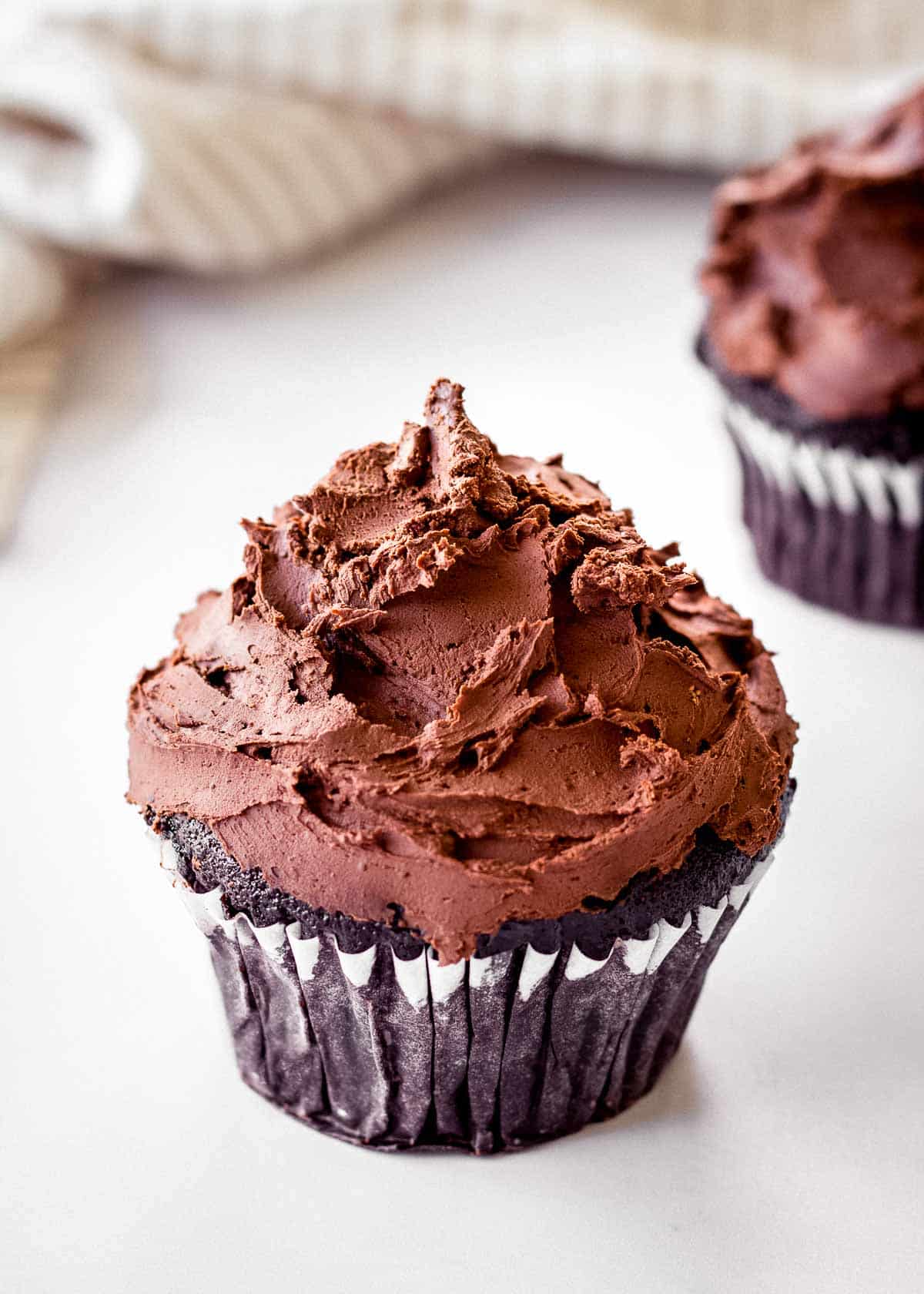 Two chocolate cupcakes decorated with vegan dark chocolate ganache. A tea towel sits in the background.