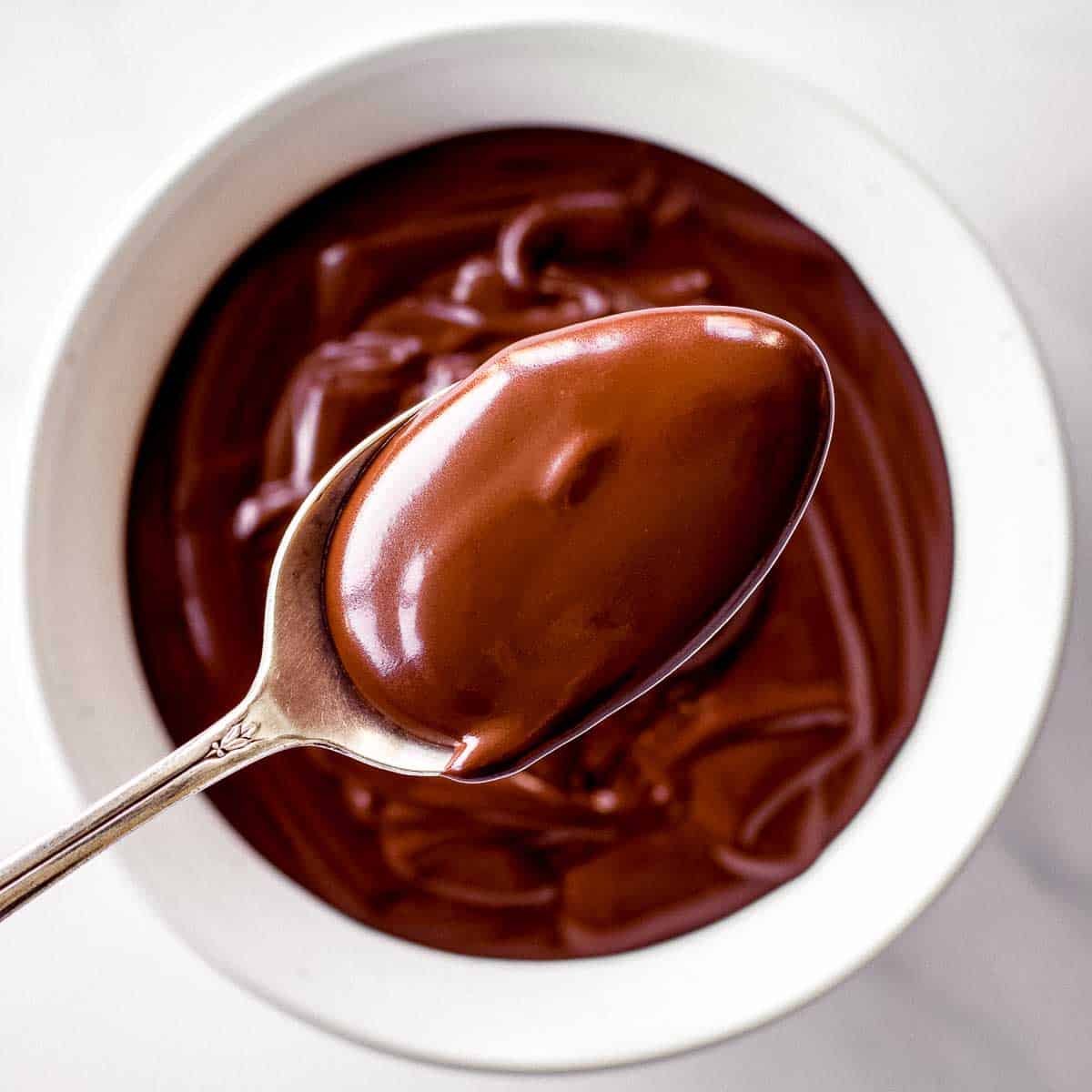 A silver spoon of vegan chocolate ganache hovers over a white bowl full of it.
