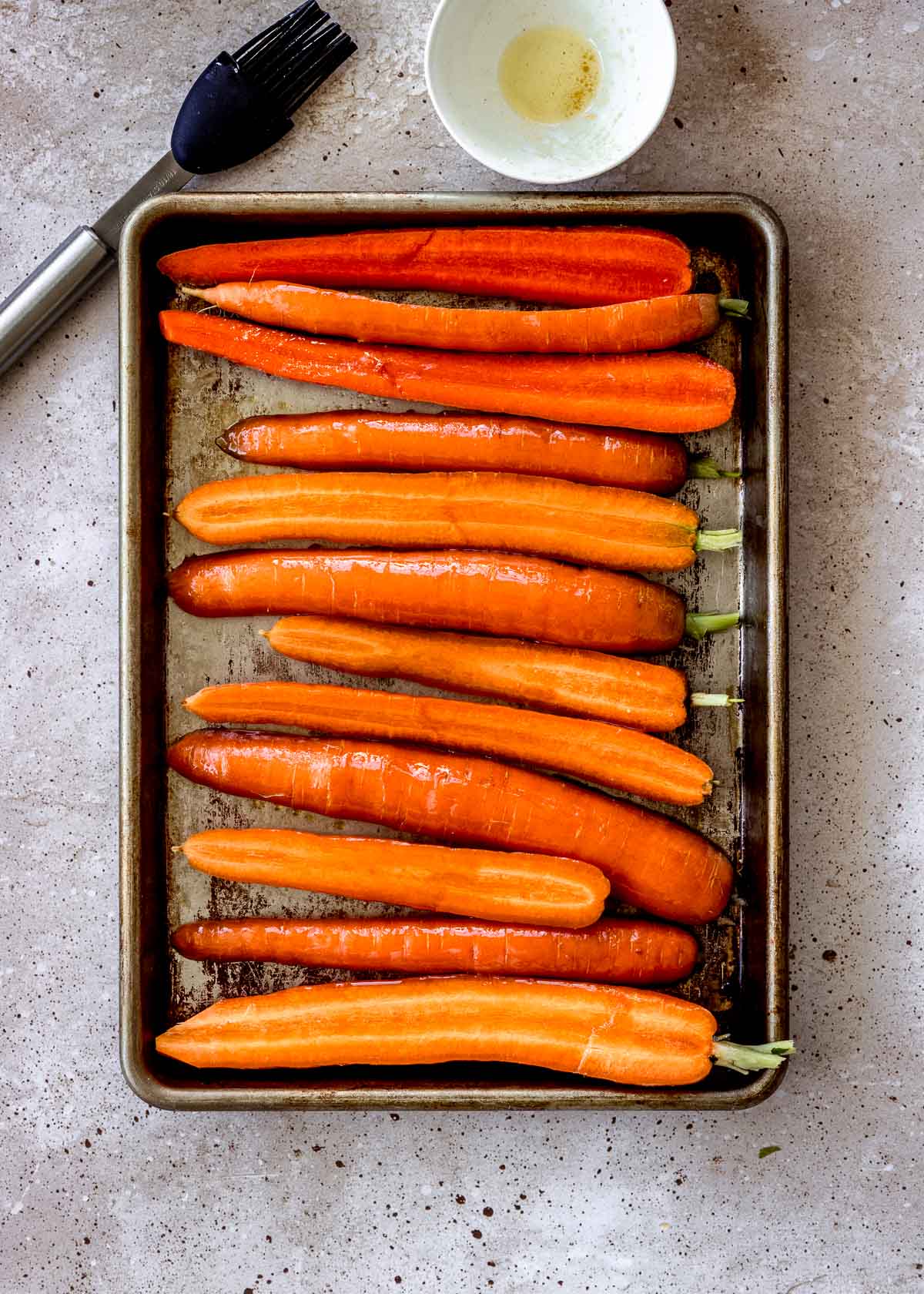 Uncooked carrots sliced in half on a baking sheet, brushed with olive oil and maple syrup. Pastry brush is nearby.