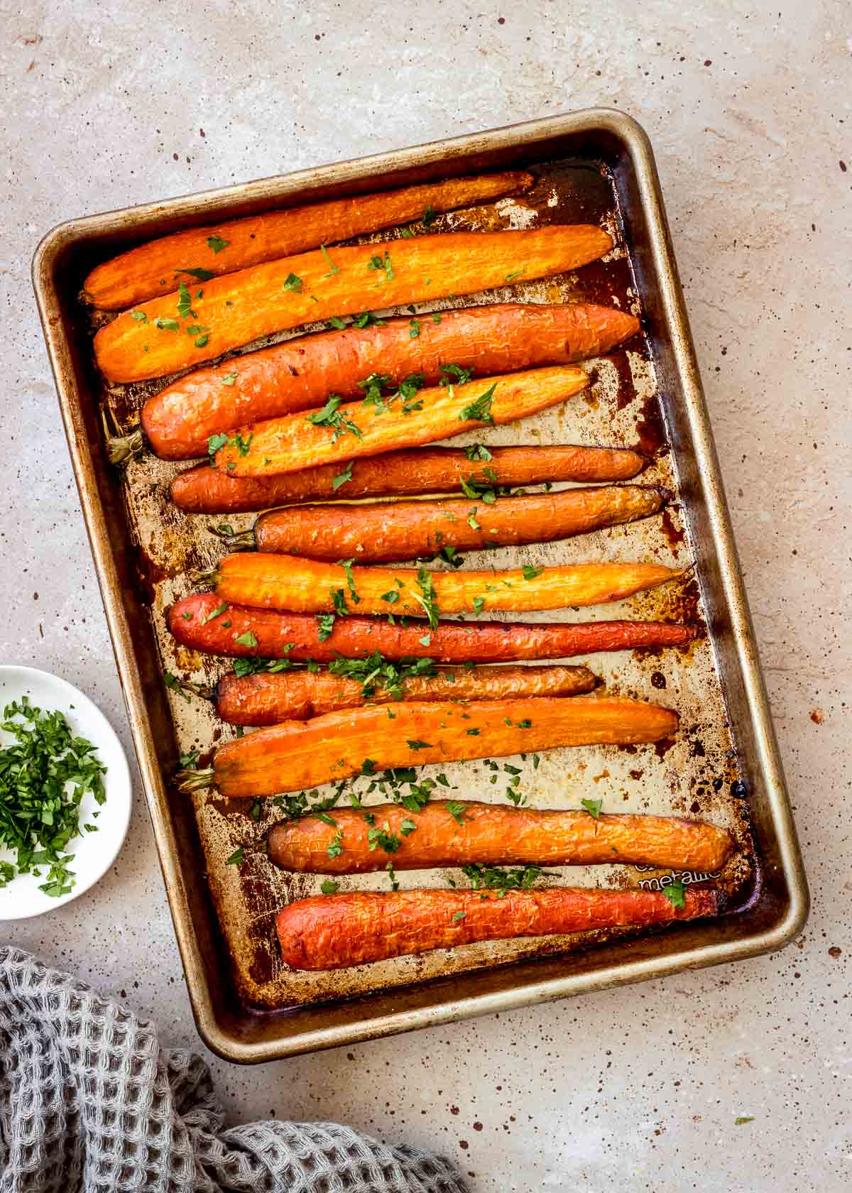 Maple roasted carrots on a silver baking sheet decorated with chopped parsley, with a dish of it nearby.