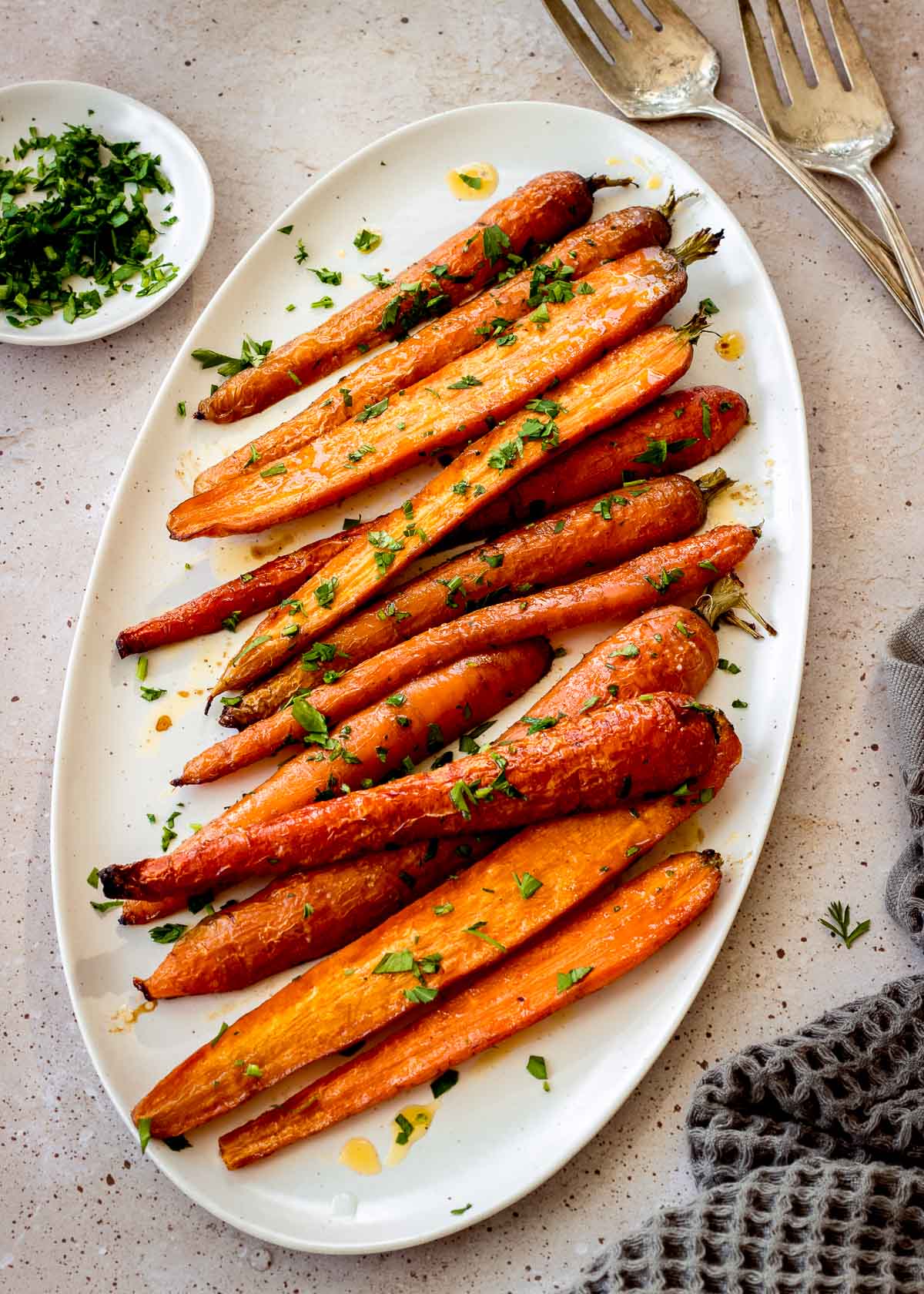 Halved maple roasted carrots on a white oblong plate, decorated with chopped parsley. Two large silver serving spoons and more parsley are nearby.