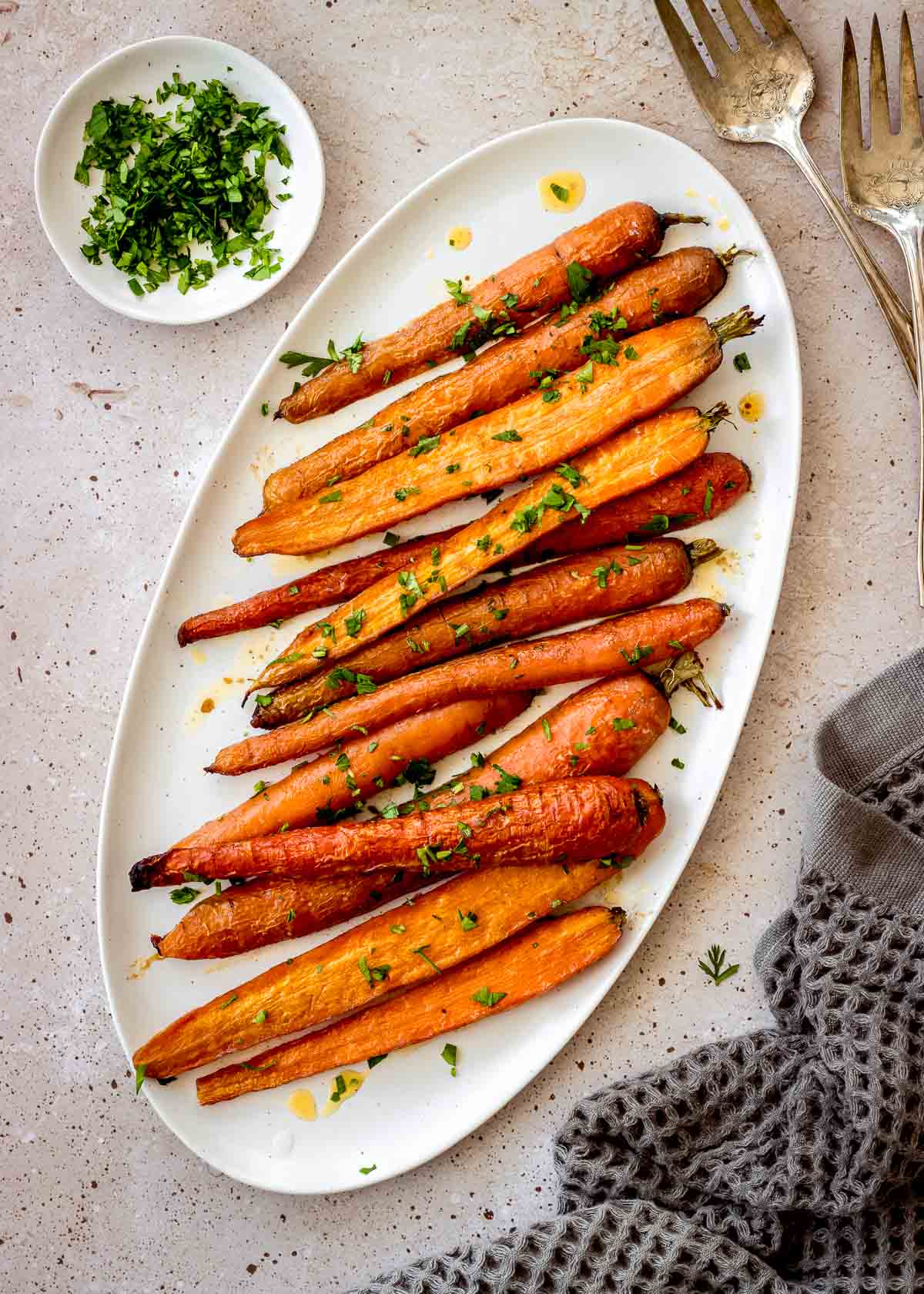 Halved roasted carrots with maple syrup sit on a white oblong plate, decorated with chopped parsley. More parsley, a tea towel and two large silver serving forks are nearby.