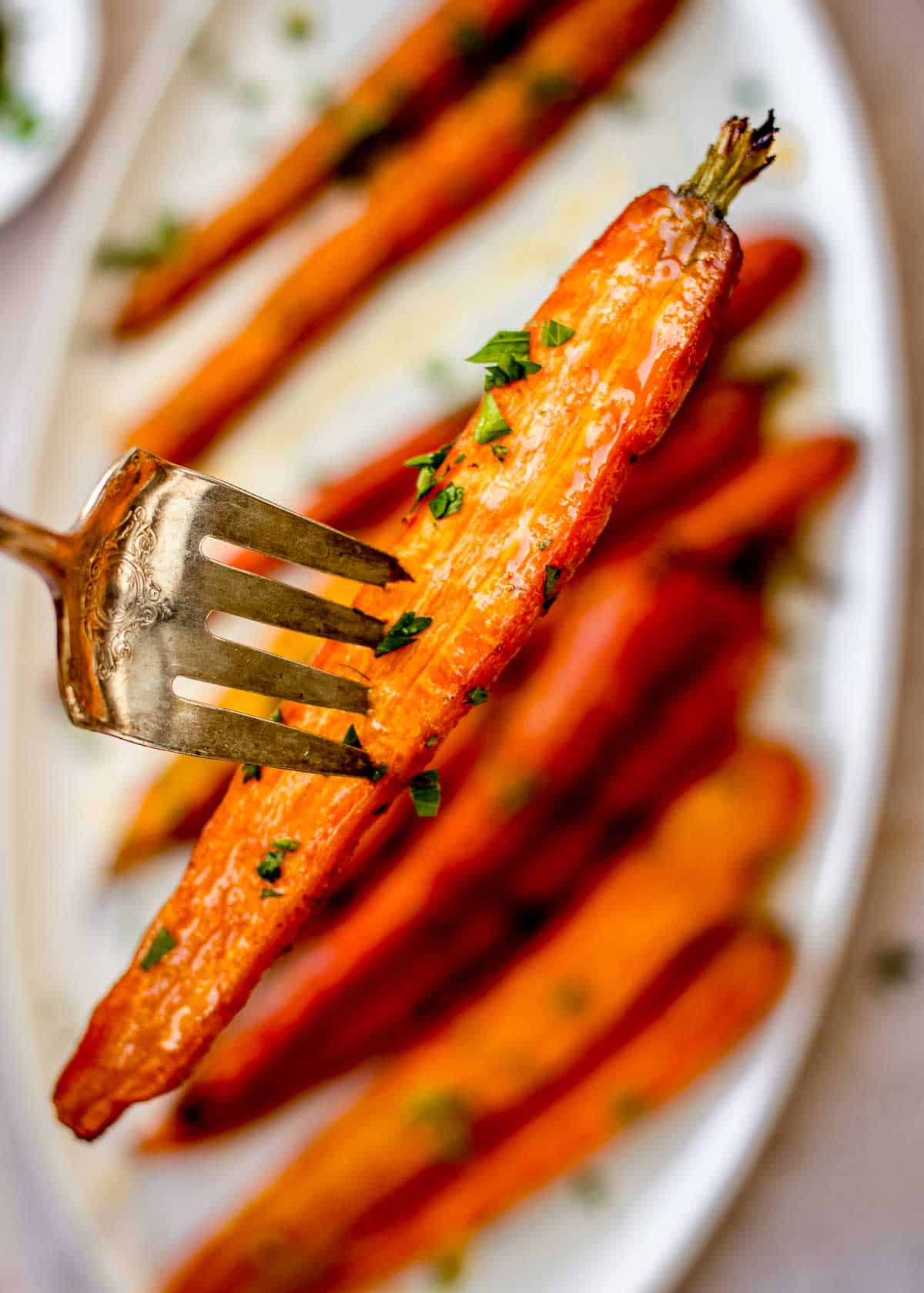 A fork with a roasted carrot on it is being held up to the camera. Behind it is a white plate of roasted carrots decorated with chopped parsley.
