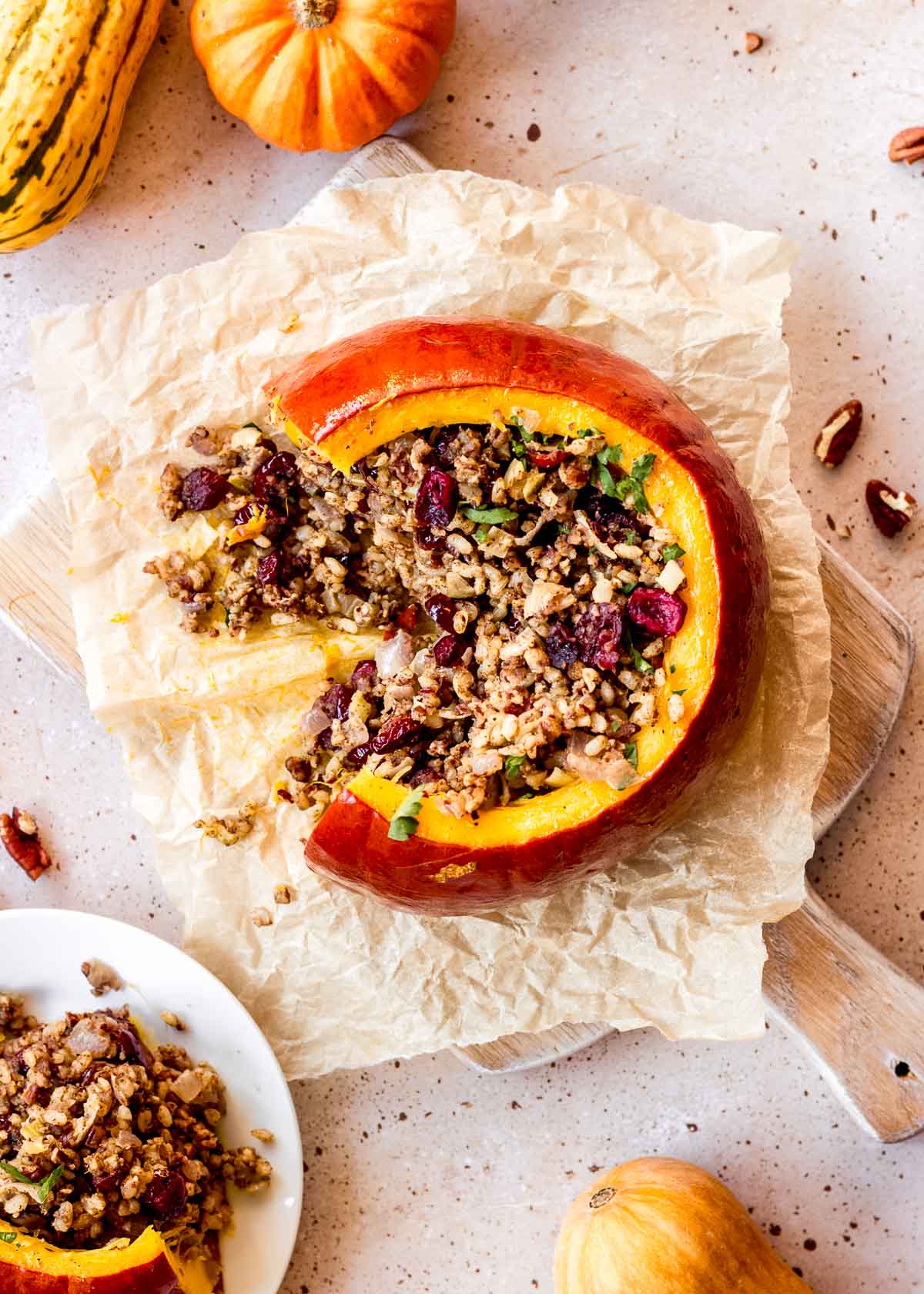 Vegan pumpkin stuffed with rice, pecans and cranberries sits on parchment paper, with small decorated squash and fork in background. A slice has been cut out of the pumpkin and it is on a plate nearby.