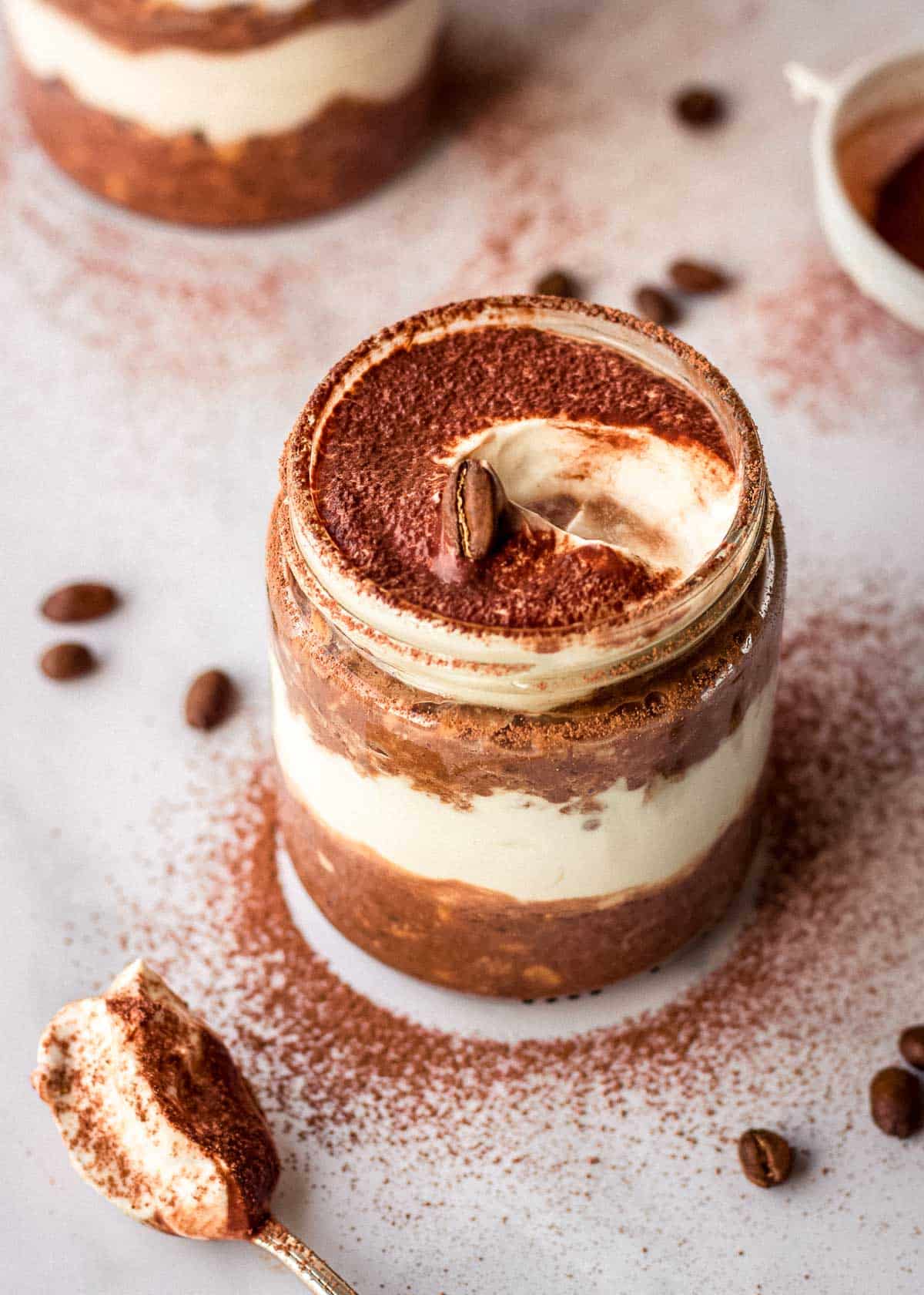Glass jar of layered tiramisu oats decorated with cocoa powder and coffee beans. A spoonful has been taken out of the jar.