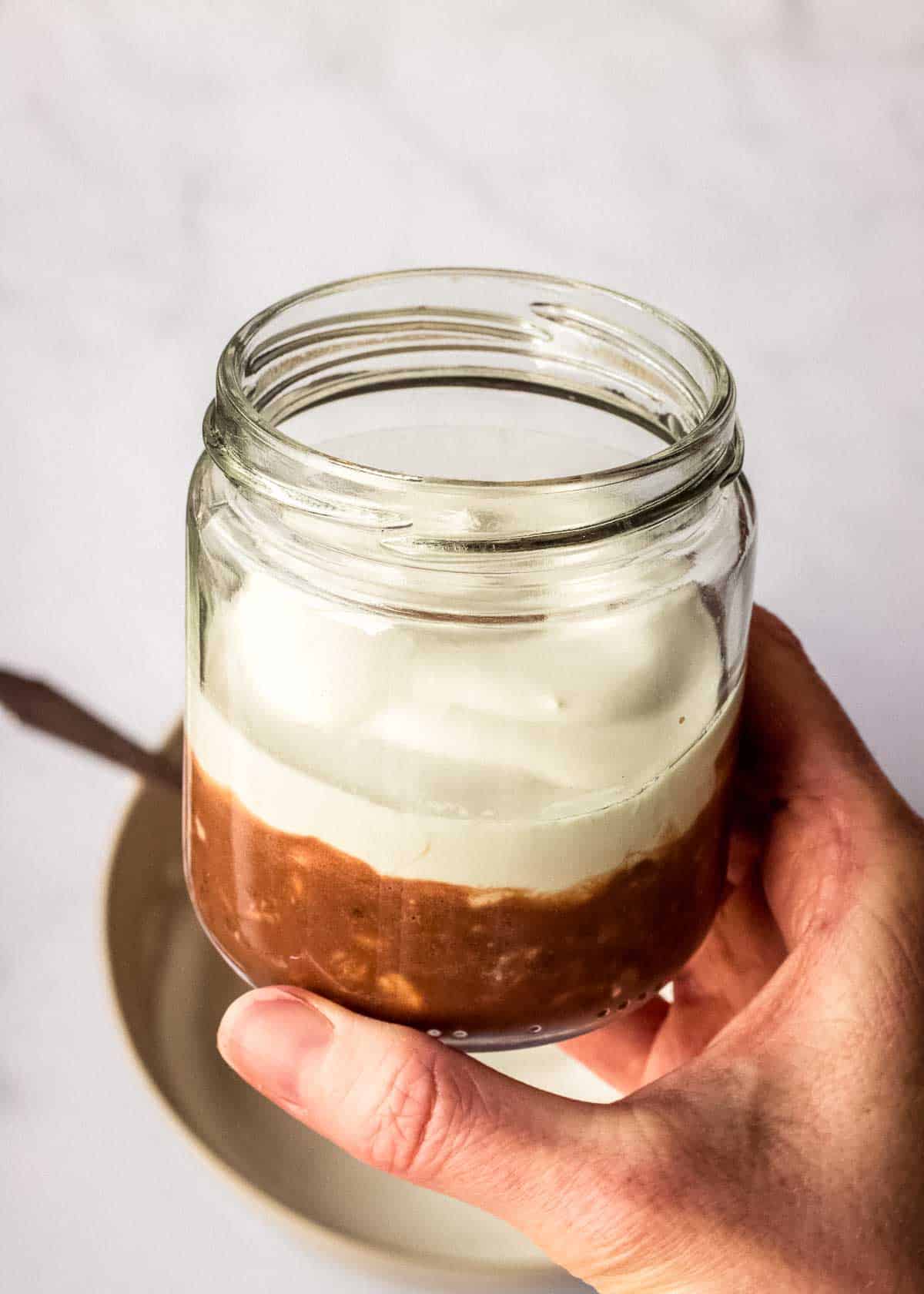 A woman holds a glass jar containing a layer of coffee chocolate overnight oats and vegan mascarpone.