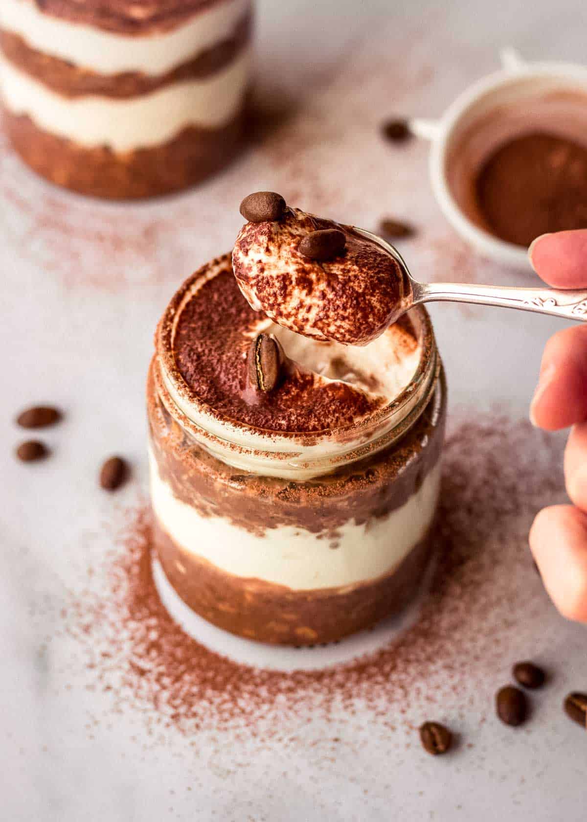 A woman takes a spoonful of tiramisu overnight oats from a glass jar, decorated with coffee beans and cacao powder.