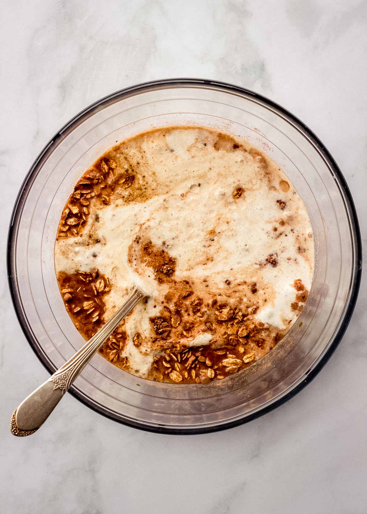 A large glass bowl of tiramisu overnight oats ingredients with a spoon in it is ready to be stirred.