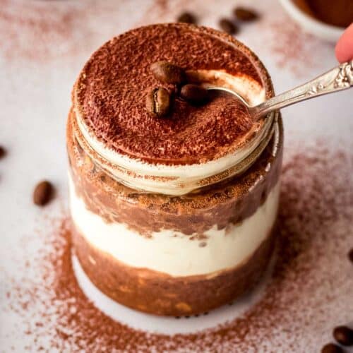 A spoonful of tiramisu overnight oats is taken from a glass jar, decorated with coffee beans and cacao powder.