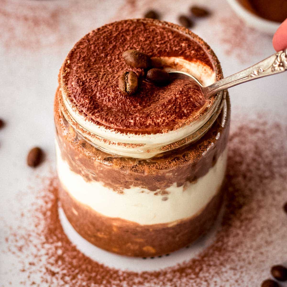 A spoonful of tiramisu overnight oats is taken from a glass jar, decorated with coffee beans and cac