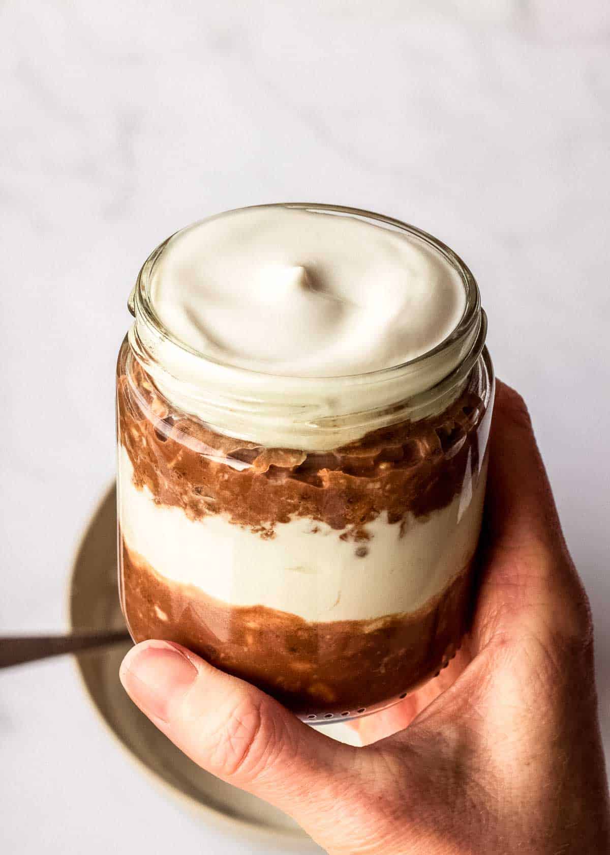 A woman holds a glass jar containing two layers of coffee chocolate overnight oats and vegan mascarpone.
