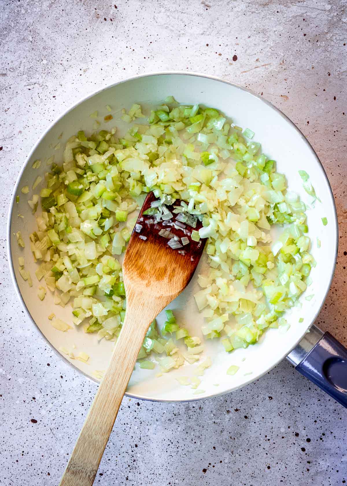 Celery, onion and garlic being sauteed in large white pan with wooden spatula.