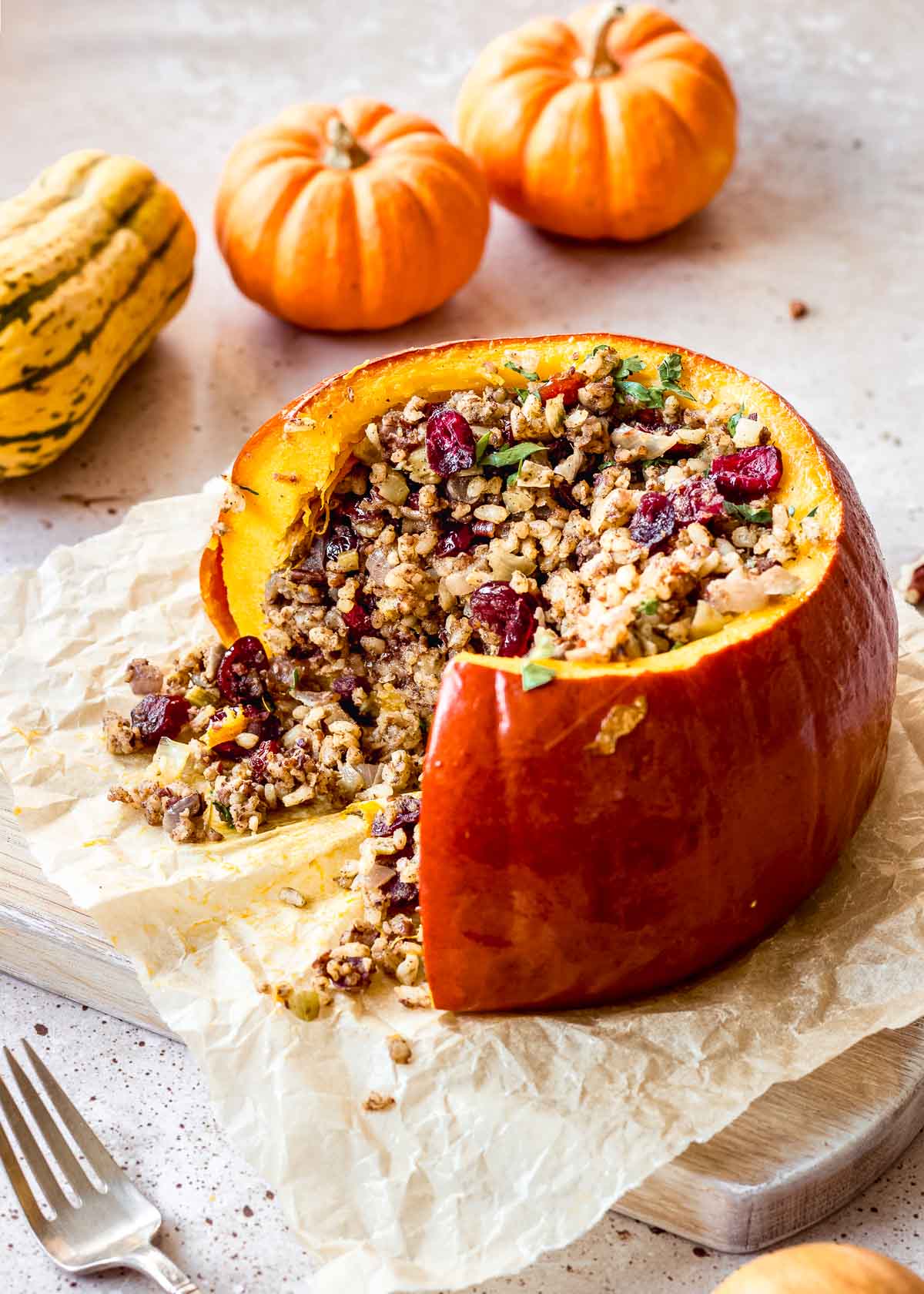 Vegan stuffed pumpkin with rice, pecans and cranberries sits on parchment paper, with small decorated squash and fork in background. A slice has been cut out of the pumpkin.