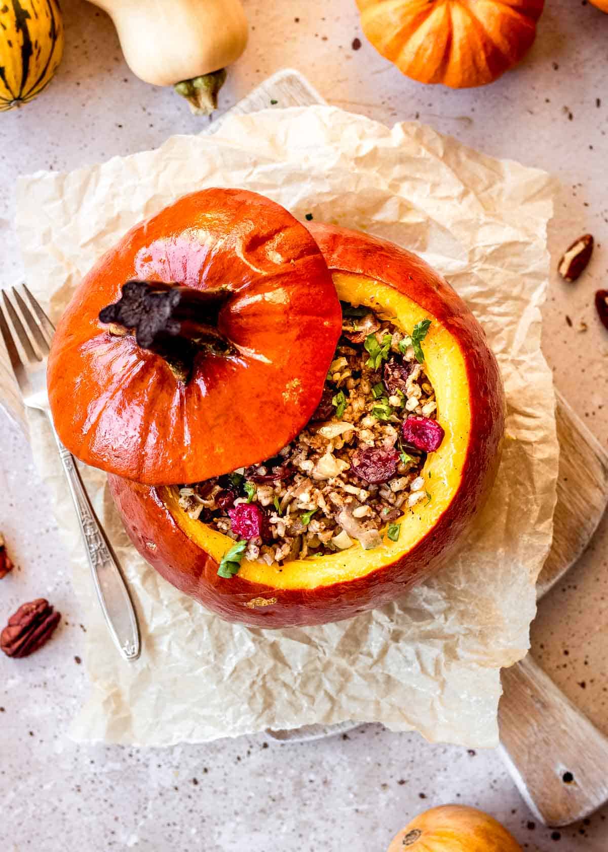 Vegan pumpkin stuffed with rice, pecans and cranberries sits on parchment paper, with it's lid on top, small decorated squash and fork in background.