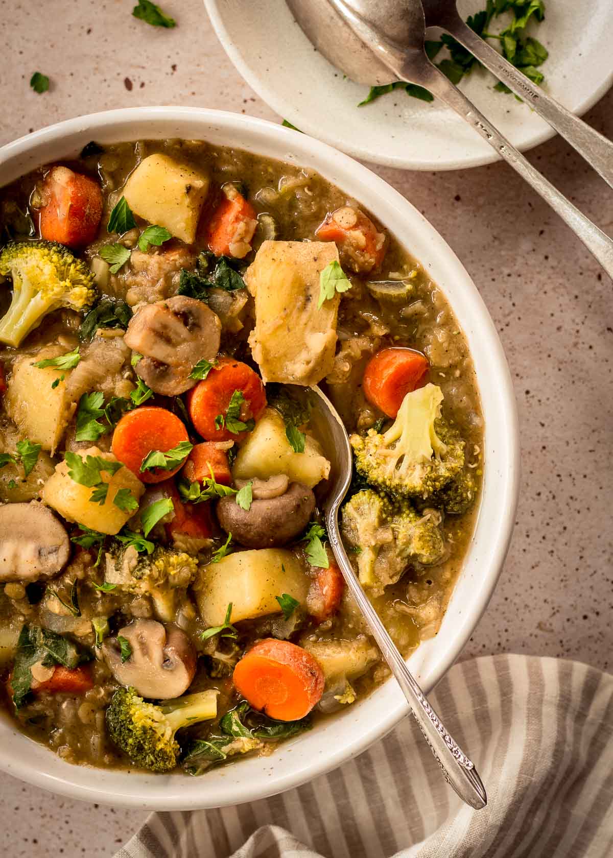 White bowl of easy vegetable stew with carrots, mushrooms, broccoli and lentils. A spoon is in the bowl and there are others nearby on a plate.