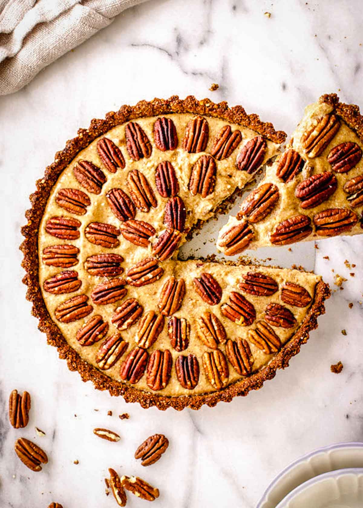 Round gluten free vegan pecan pie decorated with pecans in concentric circles, sitting on a marble background. A slice is being taken out of the pie.