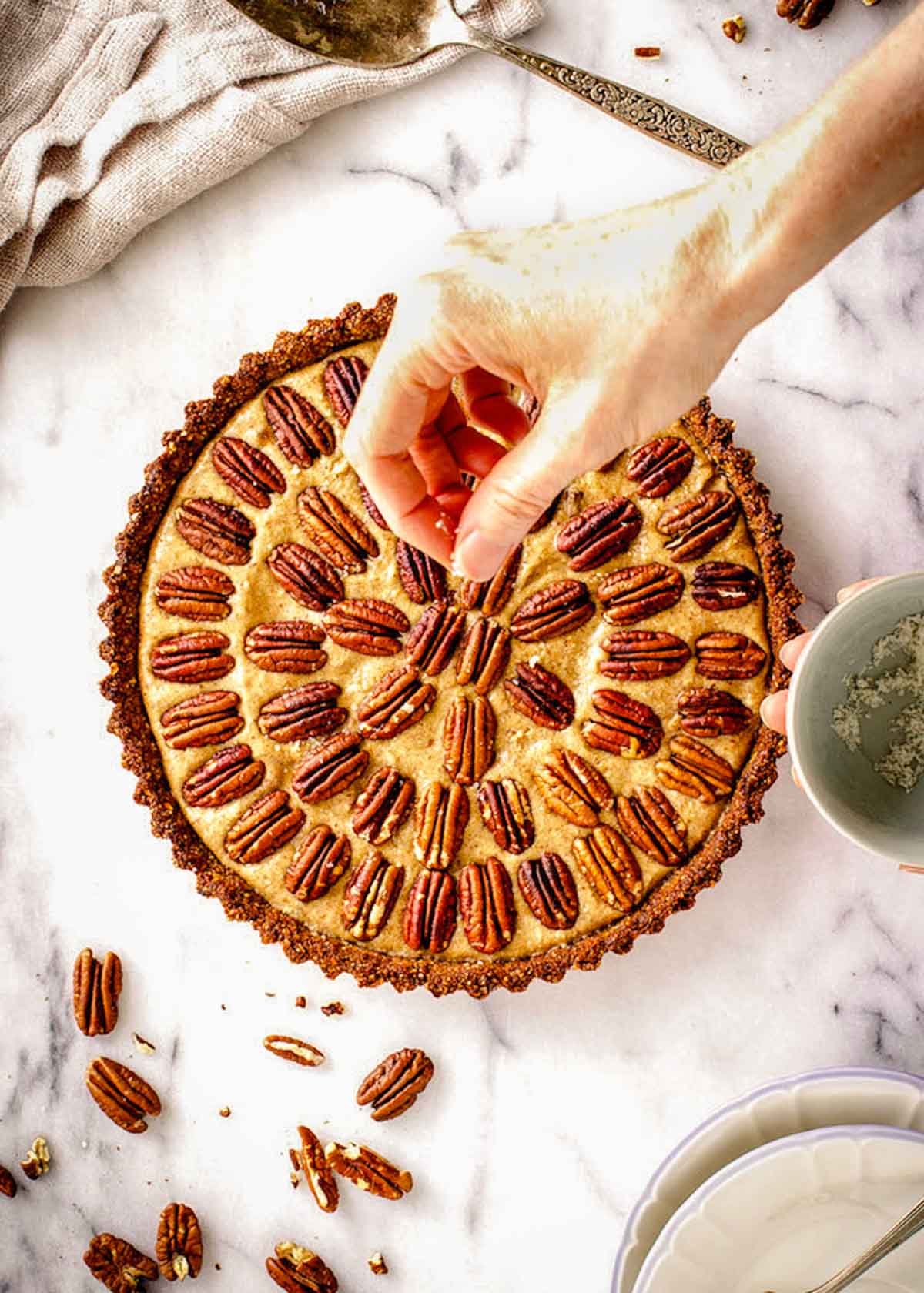 Round gluten free vegan pecan pie decorated with pecans in concentric circles, sitting on a marble background. A woman's hand is sprinkling sea salt over it.