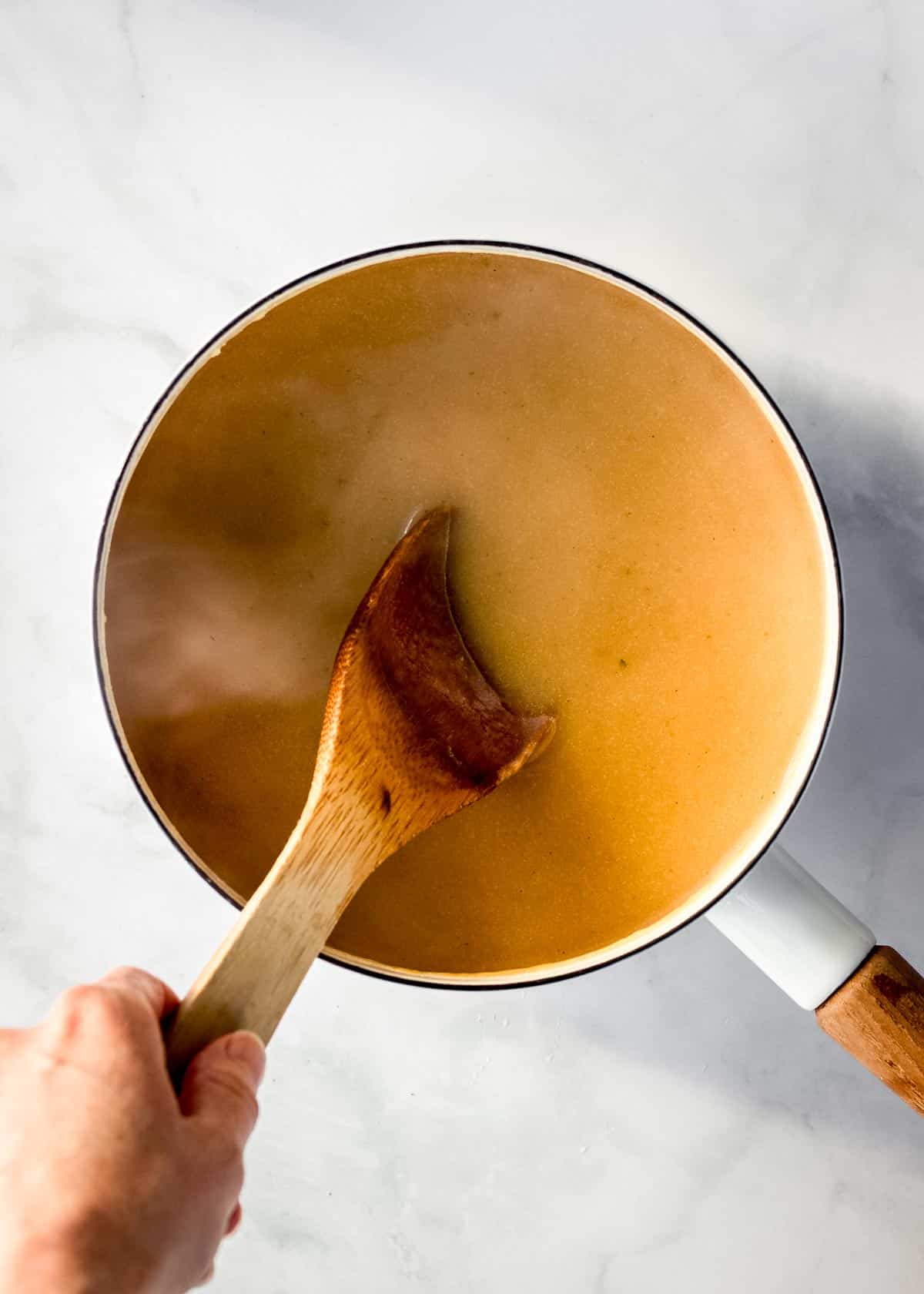 A white pot of steaming vegan gravy being stirred with a wooden spoon.