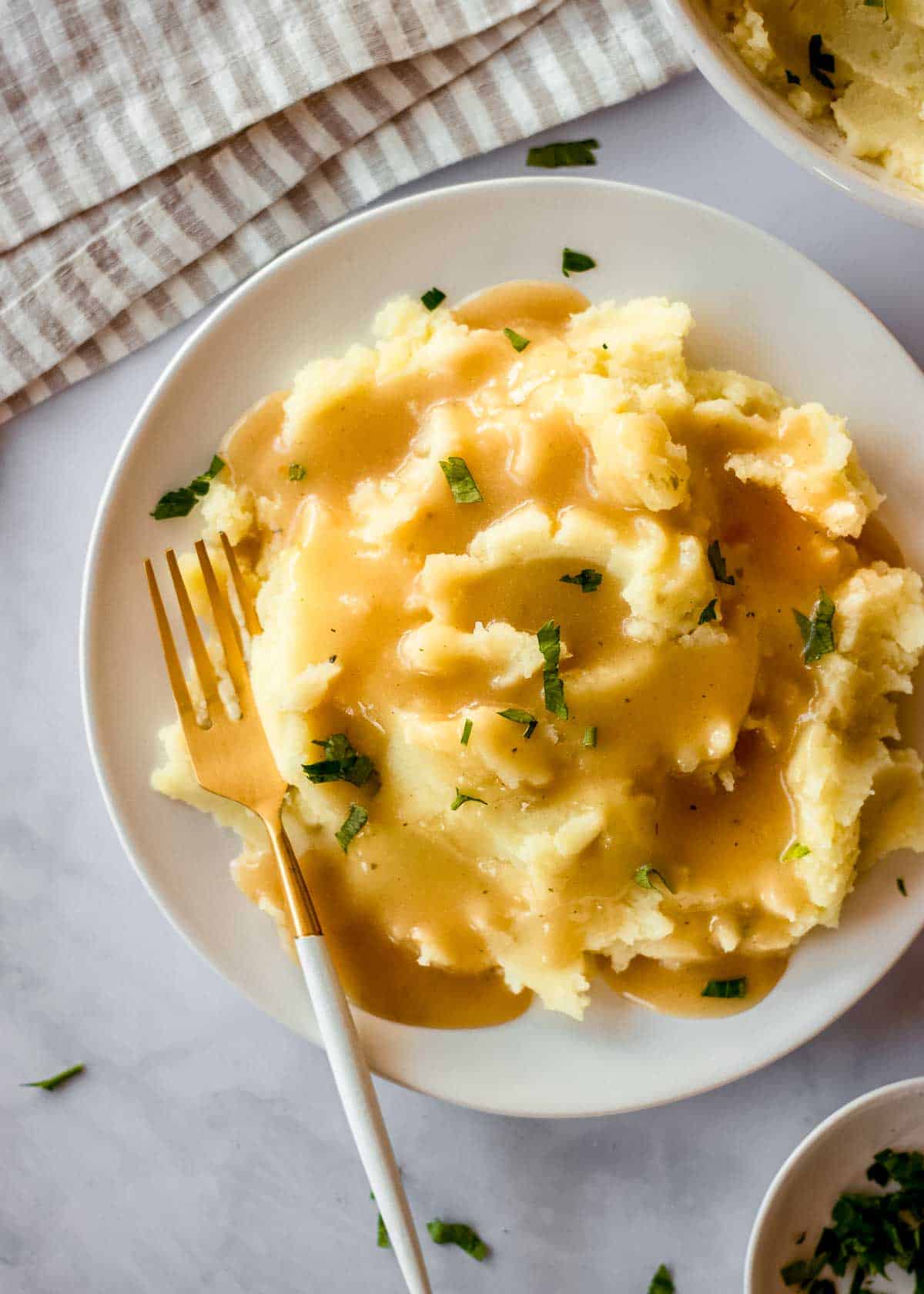 A white plate of mashed potatoes and vegan gravy sits on a white marble table. A fork is nestled in the mashed potatoes, and a dish of chopped parsley and a striped napkin are off to one side.