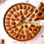 Round vegan pecan pie decorated with pecans in concentric circles, sitting on a marble background. A slice is being taken out of the pie.