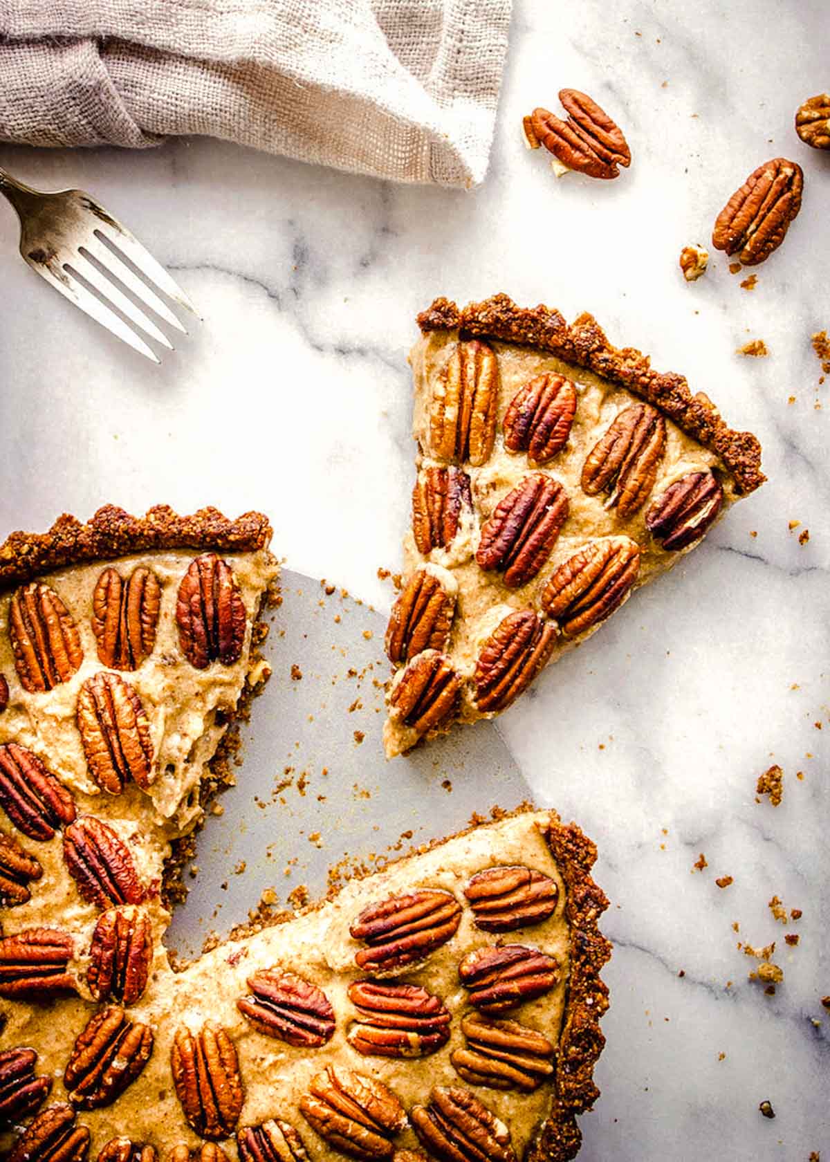 A vegan gluten free pecan pie sits on a marble counter with a slice taken out. A fork and napkin sit nearby.