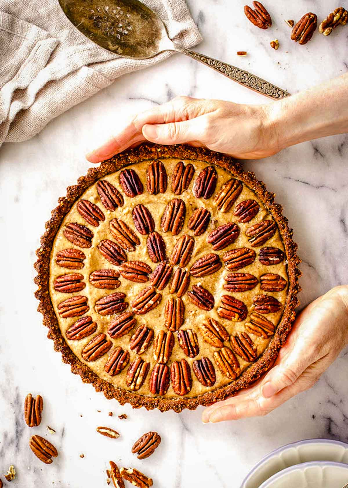 A woman's hands hold a vegan pecan pie decorated with concentric circles of pecans. A silver cake slicer, napkin and places are nearby.