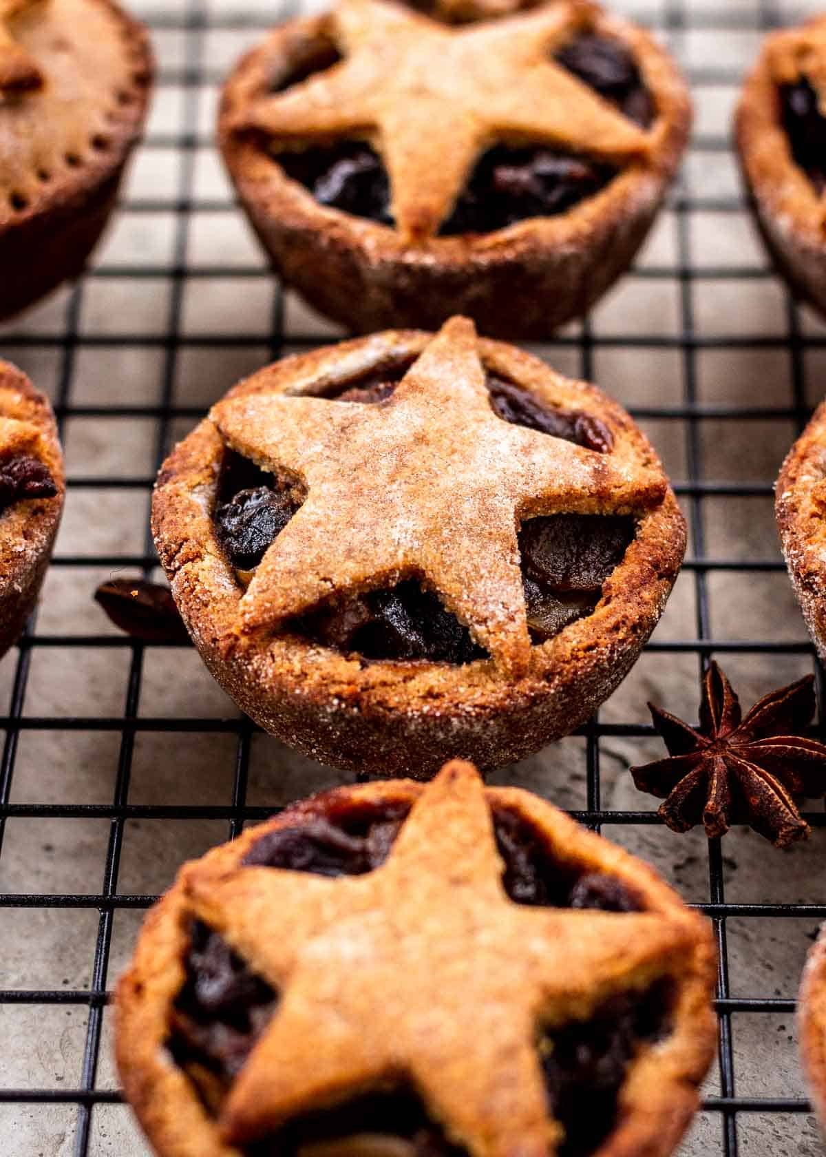 Vegan mince pies that are gluten free with pastry stars on top sit on wire cooling rack. Star anise and Christmas lights sit nearby.