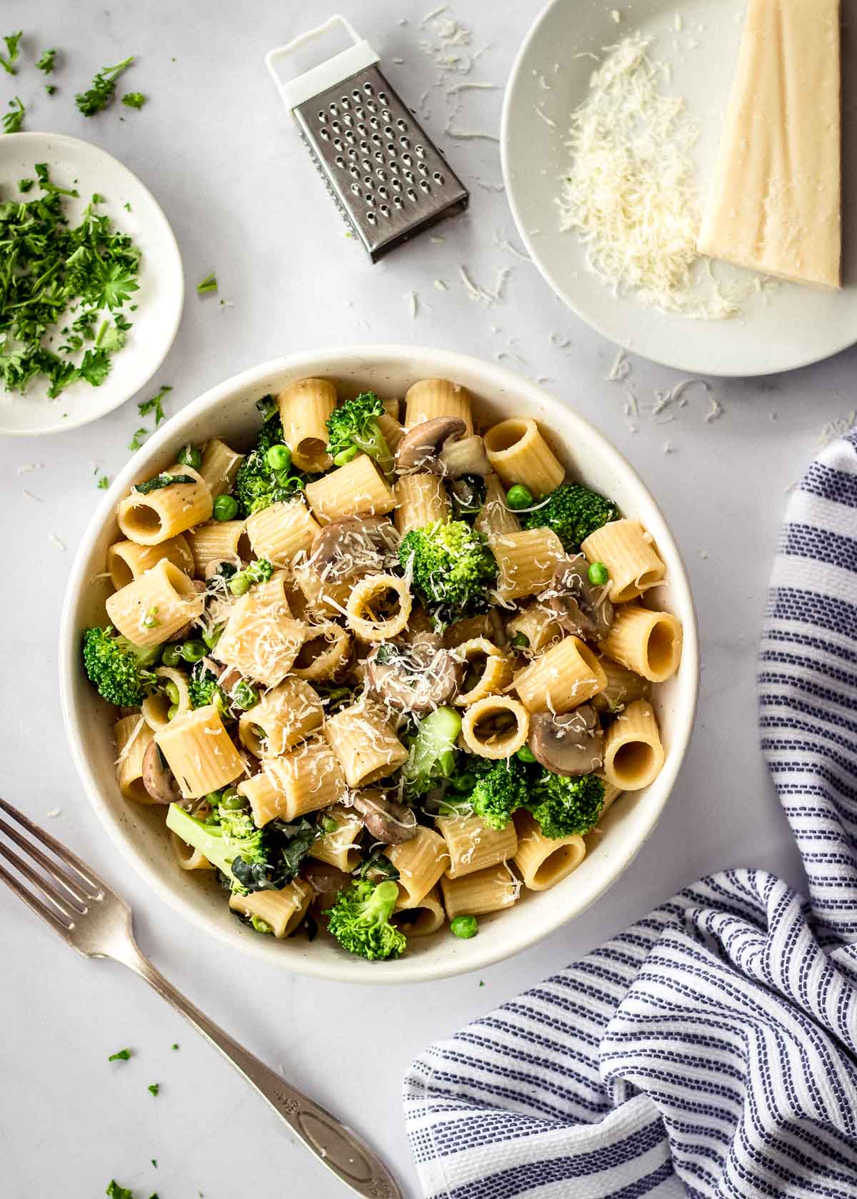 Overhead shot of bowl of broccoli mushroom pasta surrounded by vegan parmesan block and grater, a dish of parsley, a fork and napkin.