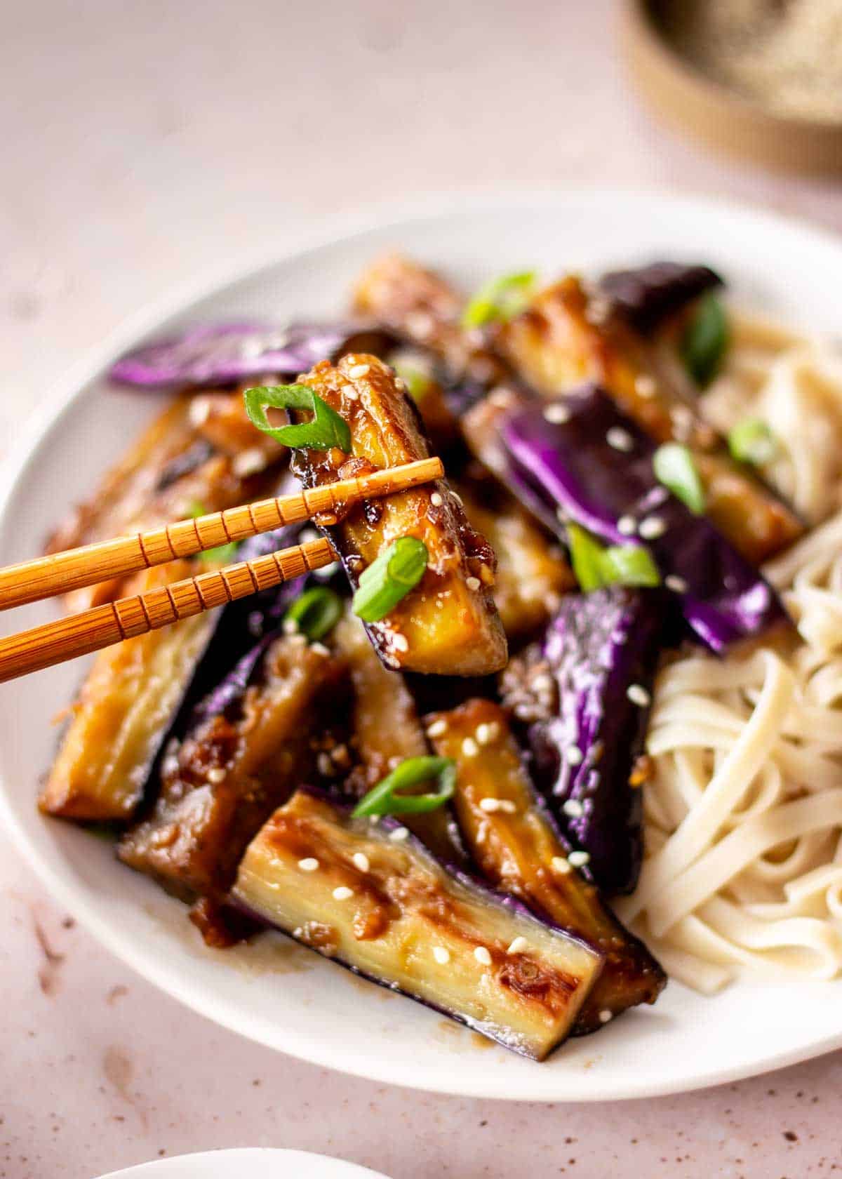 A white plate of eggplant stir fry and green onions. A piece of eggplant is being held up to the camera by wooden chopsticks.