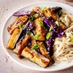 A white plate of eggplant stir fry and flat noodles, decorated with green onions and sesame seeds.
