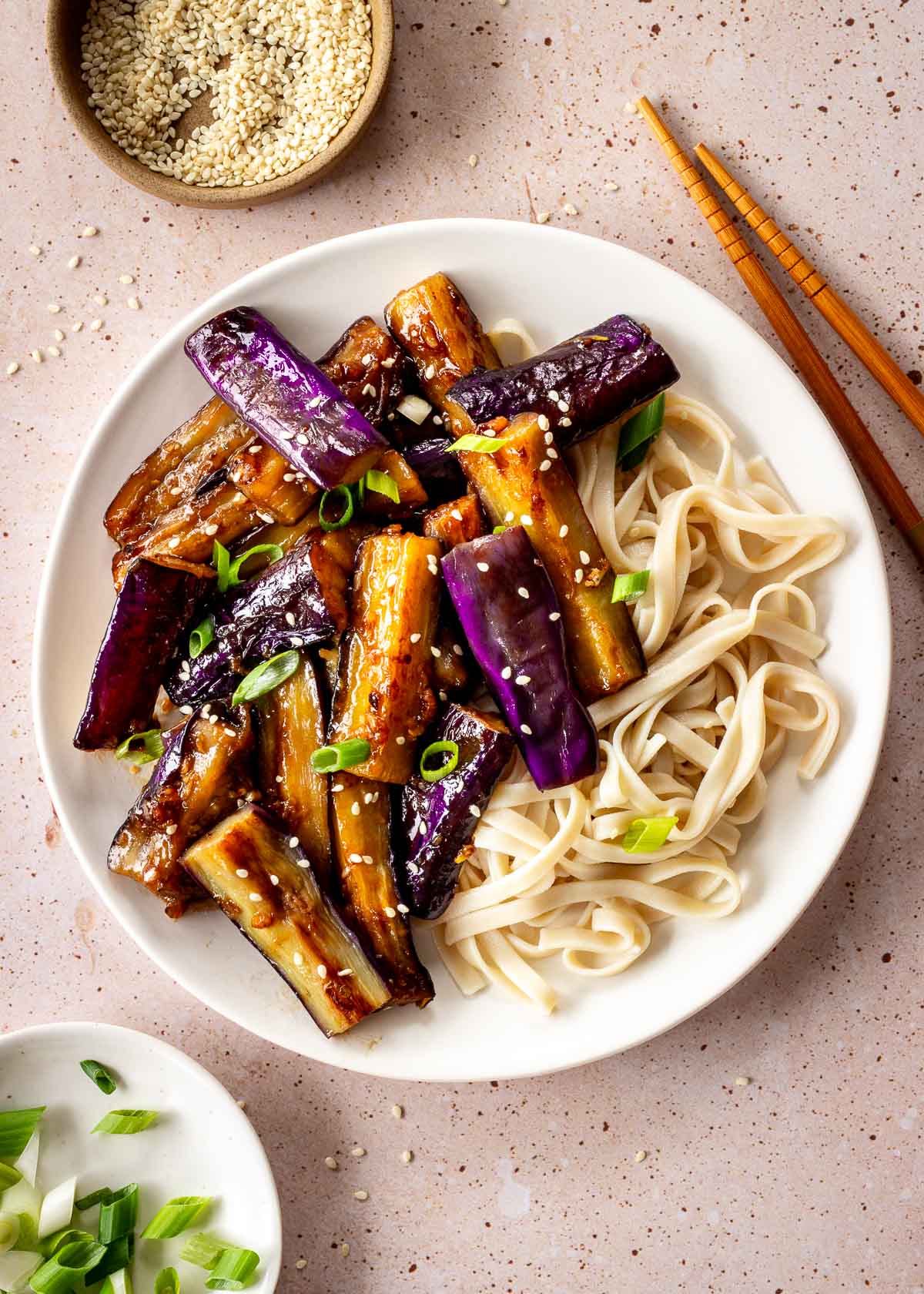 A white plate of eggplant stir fry and flat noodles, decorated with green onions and sesame seeds. A pair of chopsticks and dishes of sesame and green onions sit nearby.
