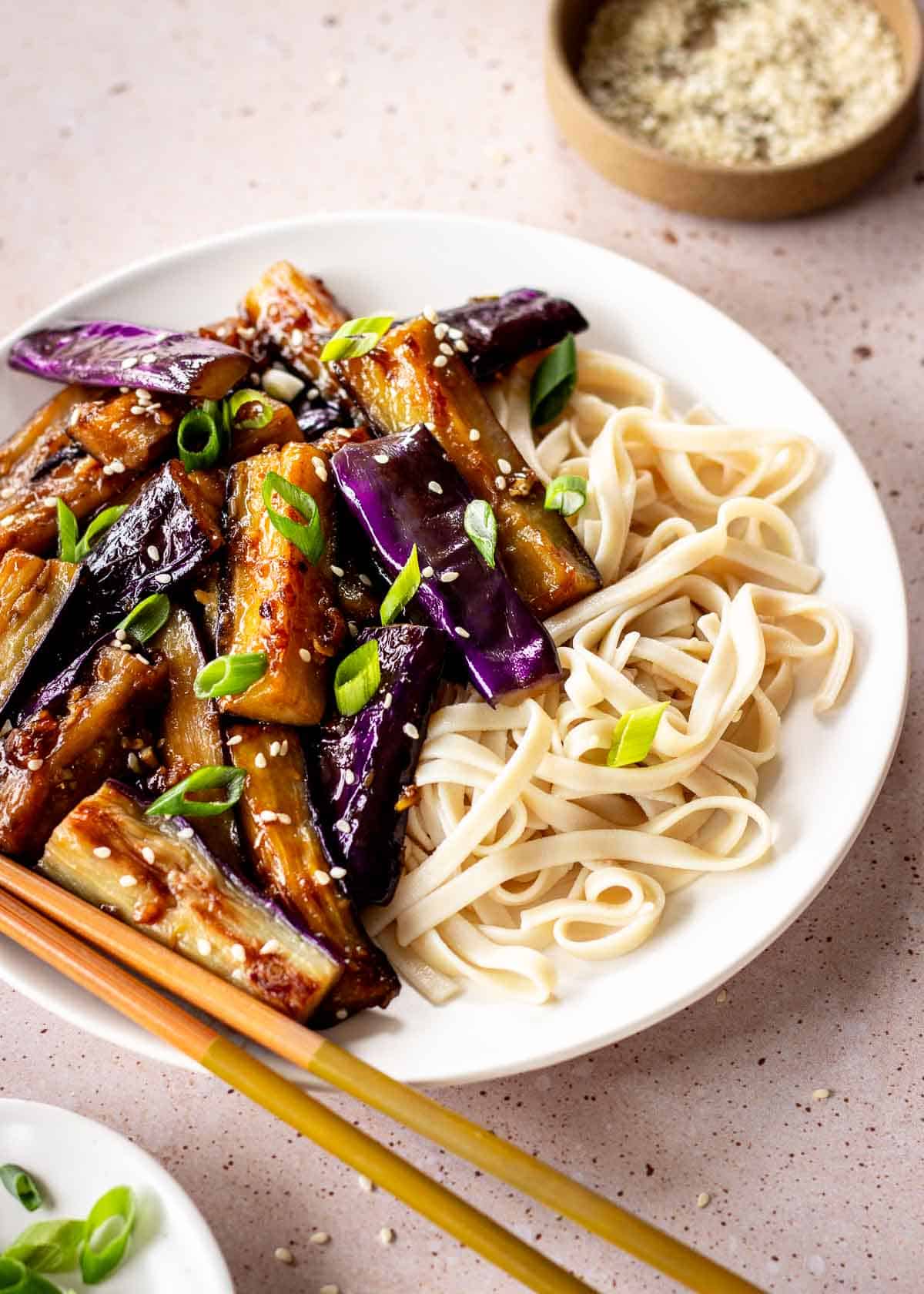 A white plate of eggplant stir fry and flat noodles, decorated with green onions and sesame seeds. A pair of chopsticks sits on the side of the plate.