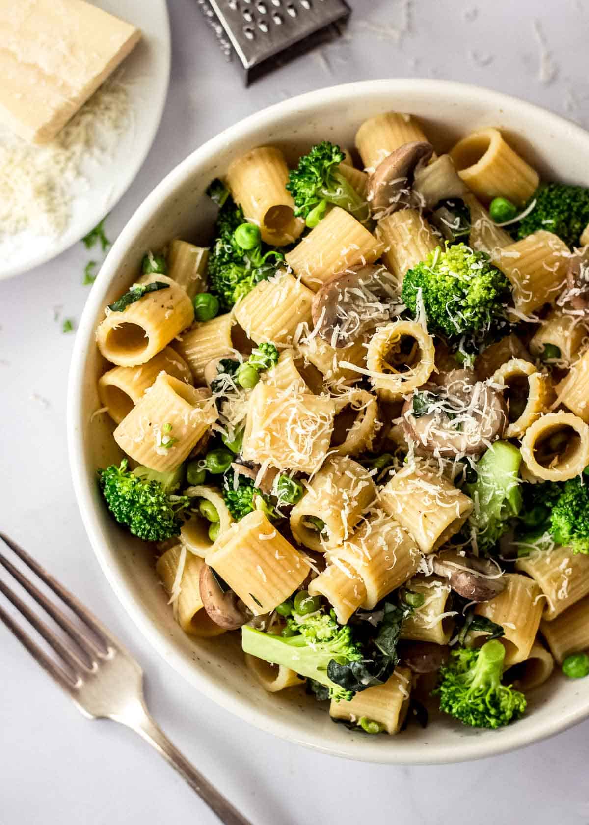 White bowl of mushroom broccoli pasta decorated with vegan parmesan, surrounded by parmesan block, grater and fork.