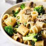 White bowl of pasta with mushrooms and broccoli, decorated with vegan parmesan.