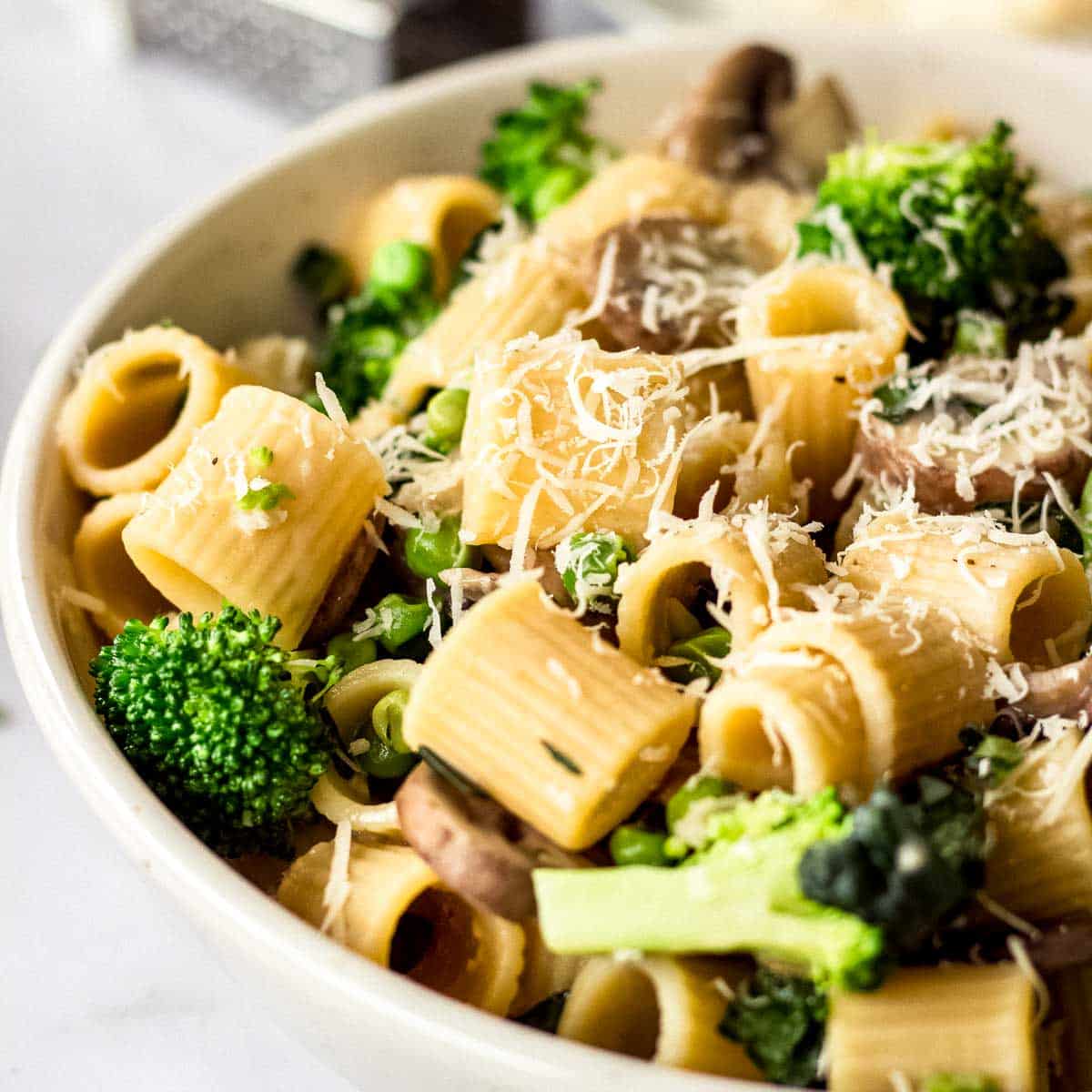 White bowl of pasta with mushrooms and broccoli, decorated with vegan parmesan cheese.
