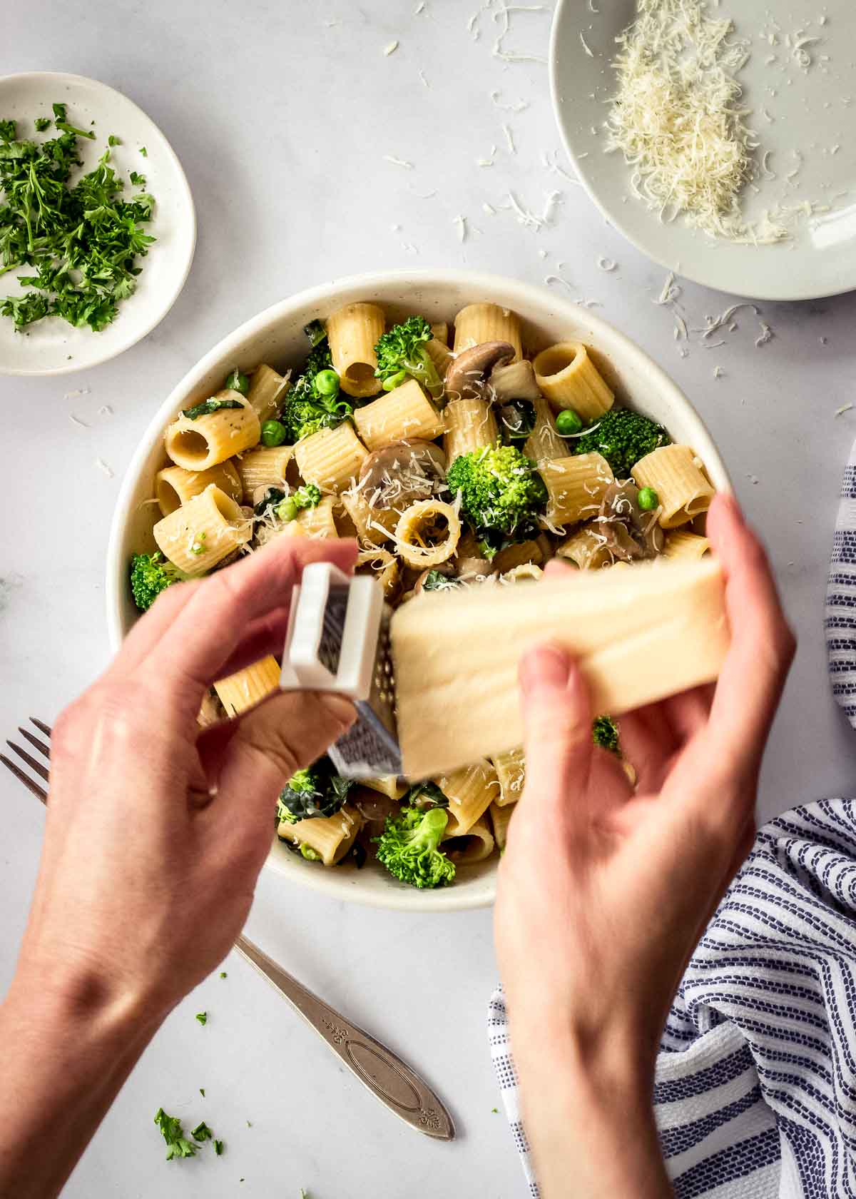 A woman's hands grate parmesan over a white bowl of pasta with mushrooms and broccoli. Dishes of parsley and parmesan surround it.