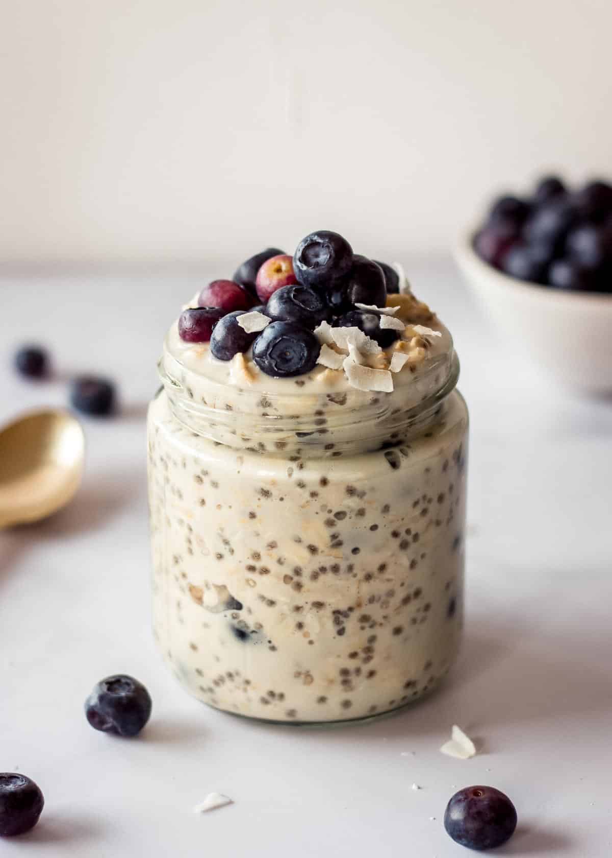 Glass jar of blueberry overnight oats topped with blueberries. There is a dish of berries in the background and a golden spoon off to one side.