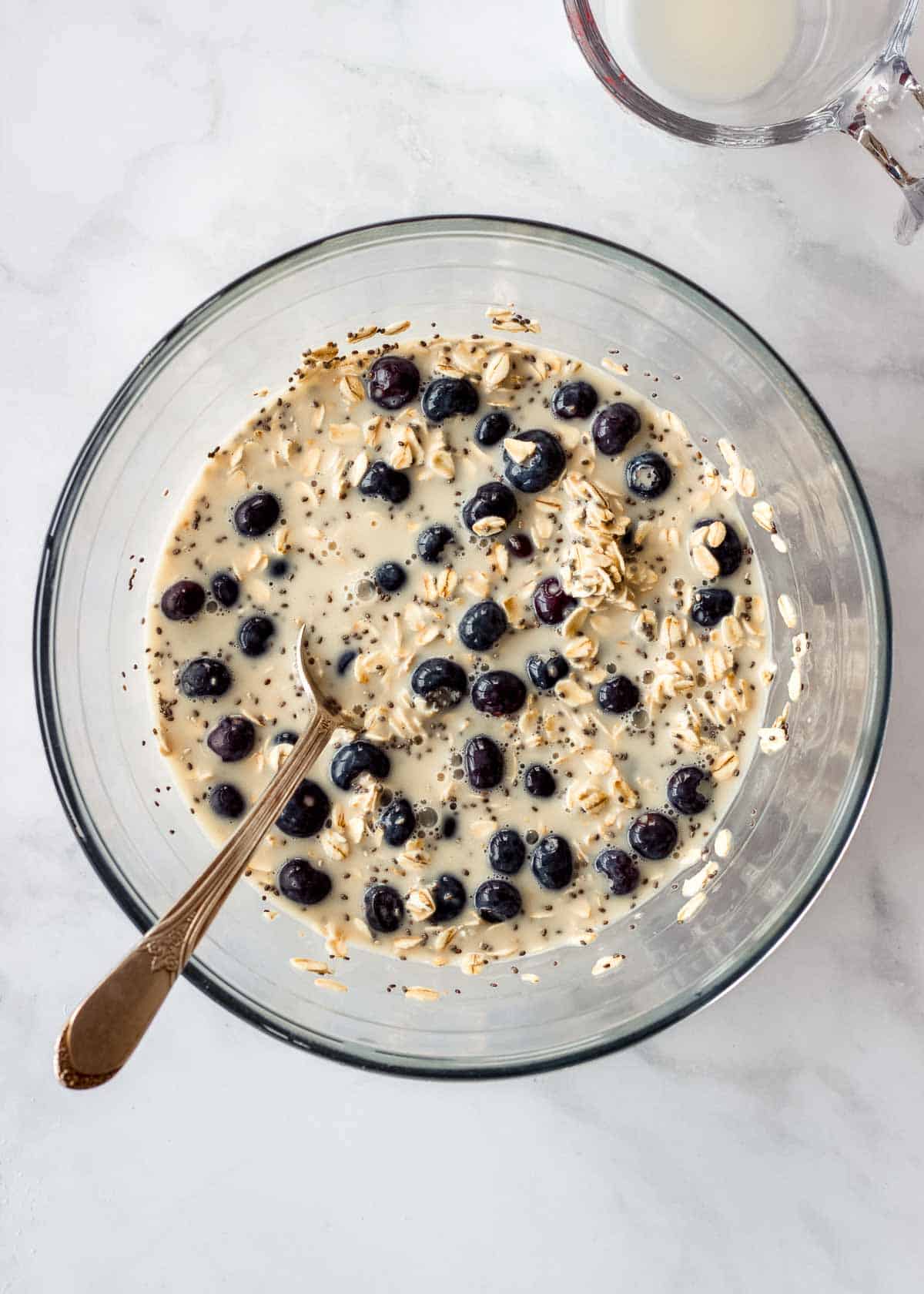 A large glass bowl of oats, blueberries, chia seeds, maple syrup and milk.