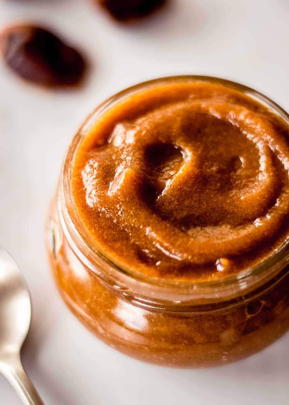 Glass jar of date paste, forming a swirl on top. There are dates in the background and a spoon nearby.