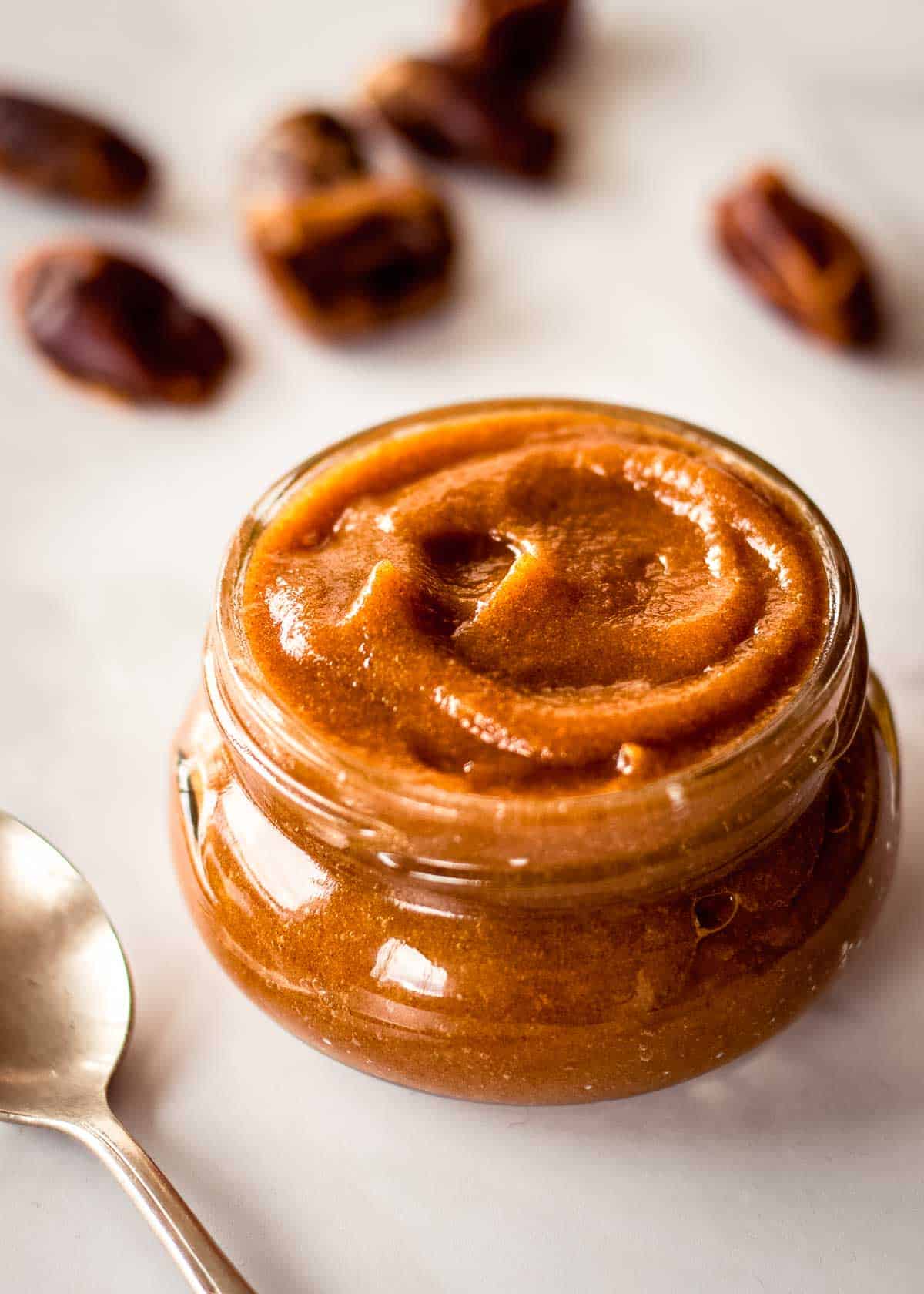 Glass jar of date puree, forming a swirl on top. There are dates in the background and a silver spoon nearby.