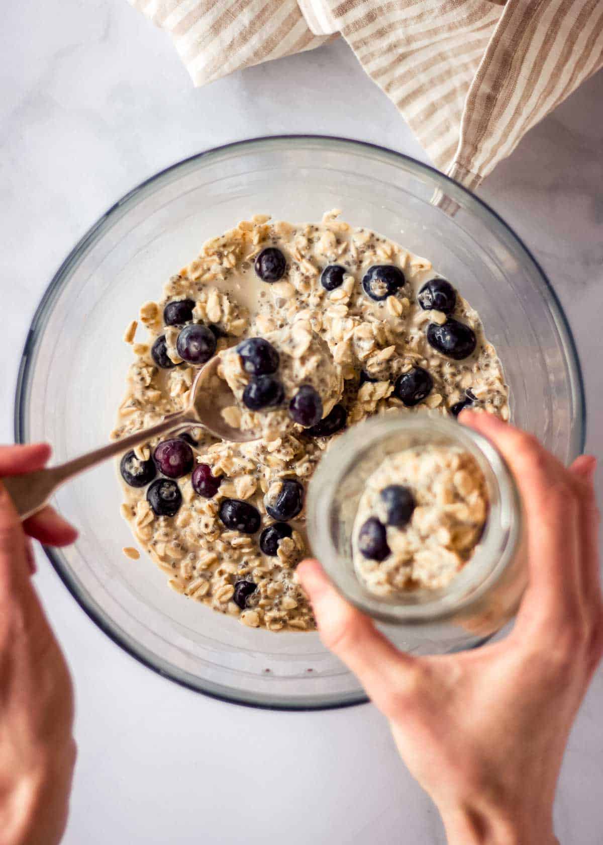 A woman's hands spoon overnight oats with blueberry from a large mixing bowl into a glass jar.
