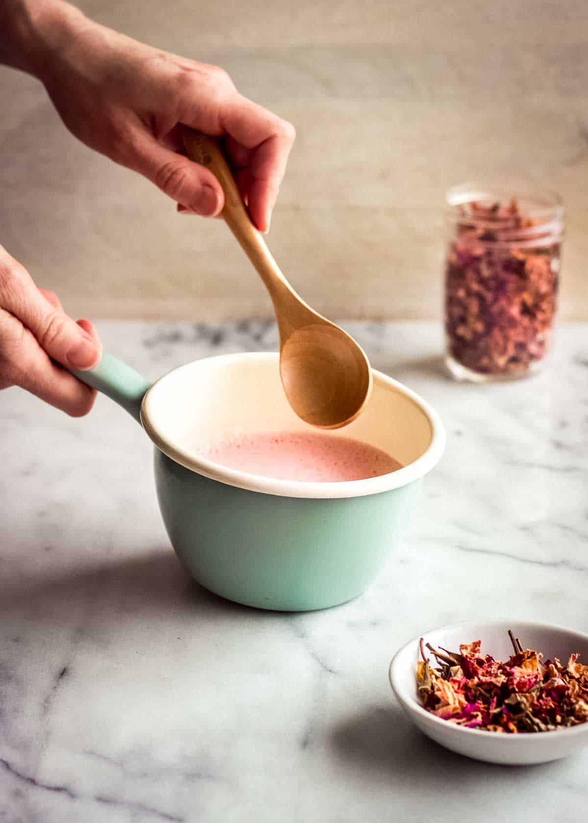 A woman's hand is stirring rose latte mix in a turquoise pan.